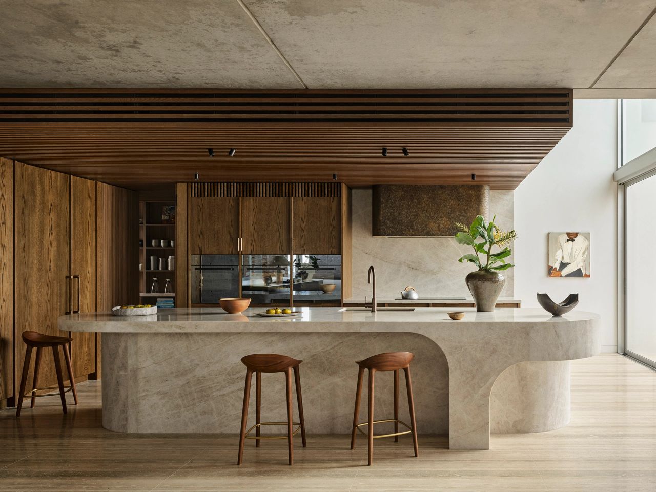 Orla contemporary kitchen blending Brazilian influences with a warm brown, black, and camel palette. Features a white and beige quartzite island with timber stools, a bronze rangehood, warm timber joinery, a large ceramic vase with palms, and timber batten detailing on the concrete ceiling.