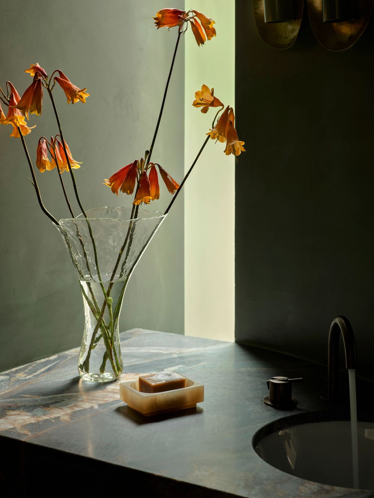Dancing on the Ceiling Textured glass vase with drooping orange bell-shaped flowers on a dark marble bathroom counter, beside a soap dish and black tapware, in moody green-toned lighting