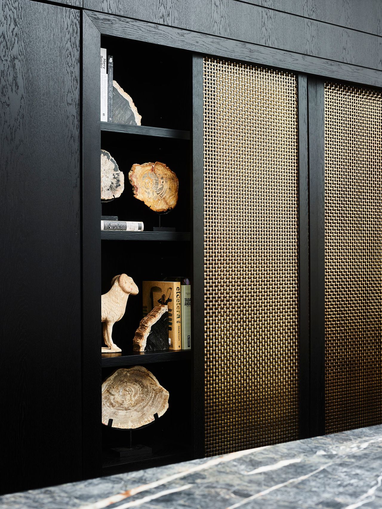 Killcare Black timber shelving unit displaying petrified wood specimens, a carved animal figurine, and books, beside sliding cabinet doors with a woven brass mesh finish