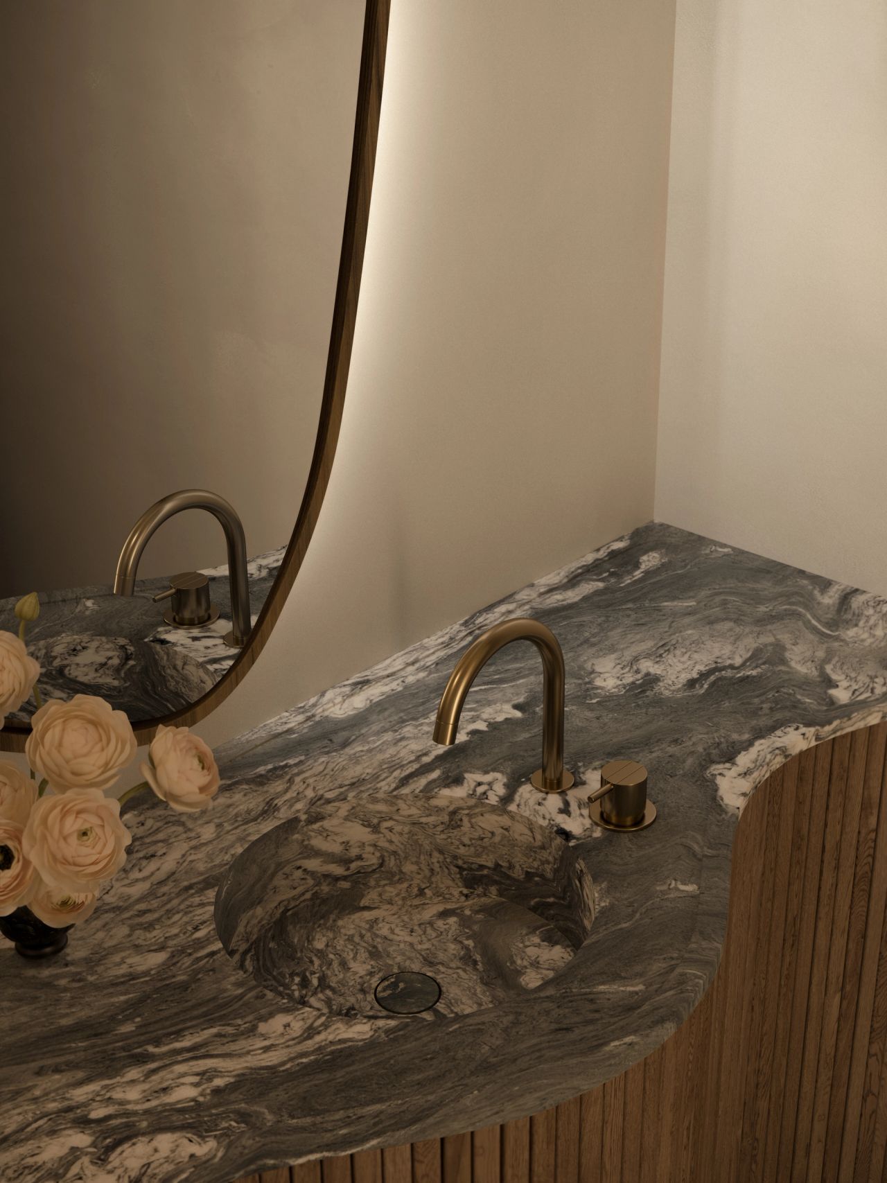 Lady M Close-up of a powder room vanity carved from dramatic grey-and-white veined stone, with an integrated sink basin and an antique brass gooseneck tap. A curved wooden-framed mirror and fluted timber frame the scene, with soft pink ranunculus in the foreground.