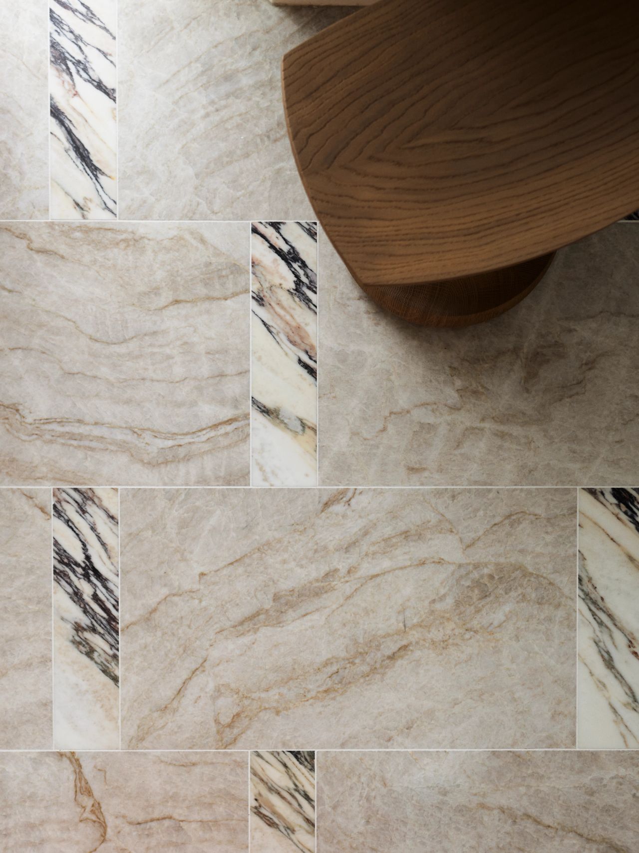 Dancing On The Ceiling Primary Ensuite Tile Detail Close-up of a beige marble tile floor with contrasting dark-veined marble accent strips, alongside the curved wooden base of a stool