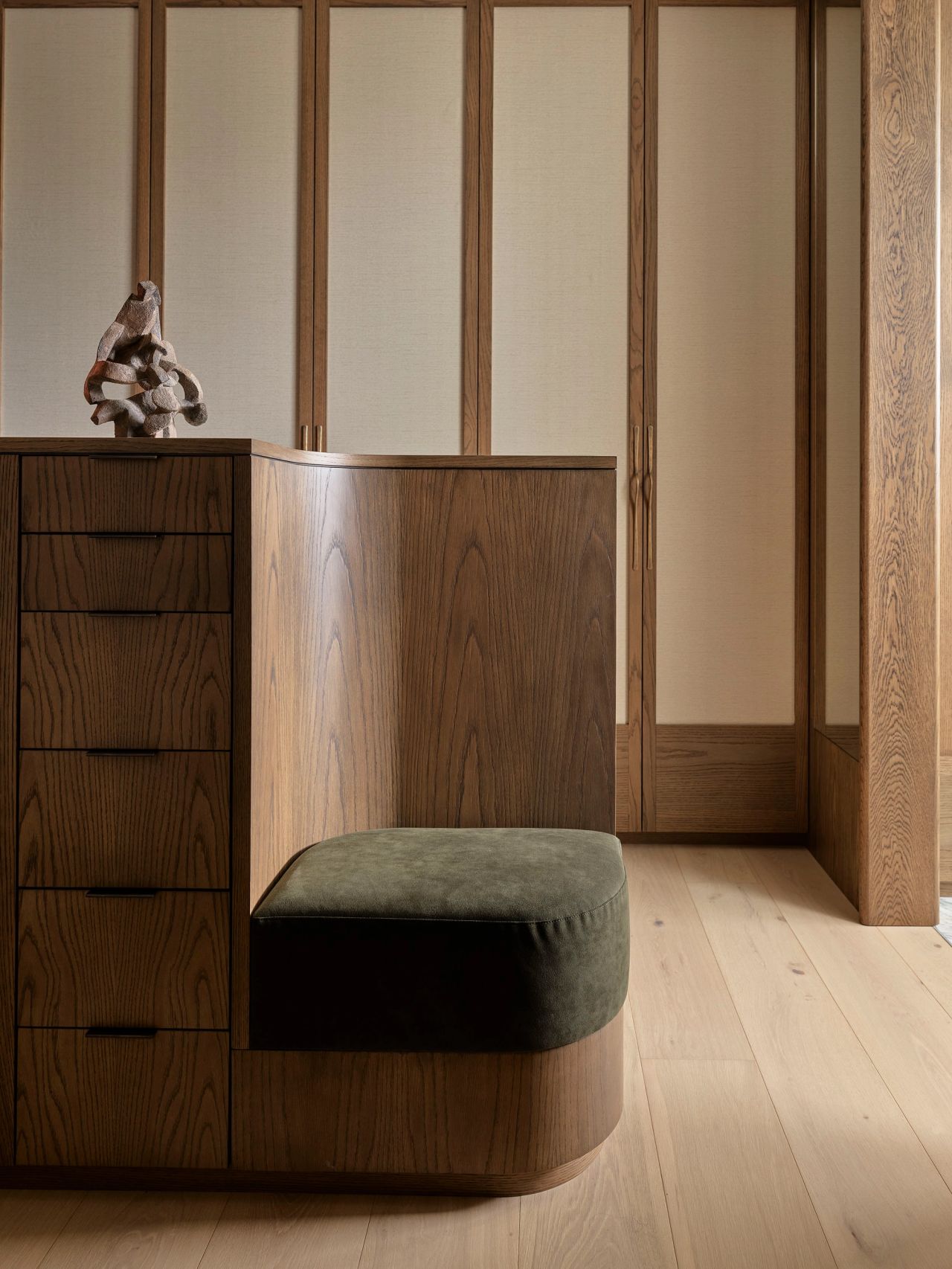 Patchwork Built-in wooden bench seat with a rounded olive velvet cushion beside a chest of drawers topped with a sculptural ceramic object, set against panelled walls in a wardrobe or dressing area.