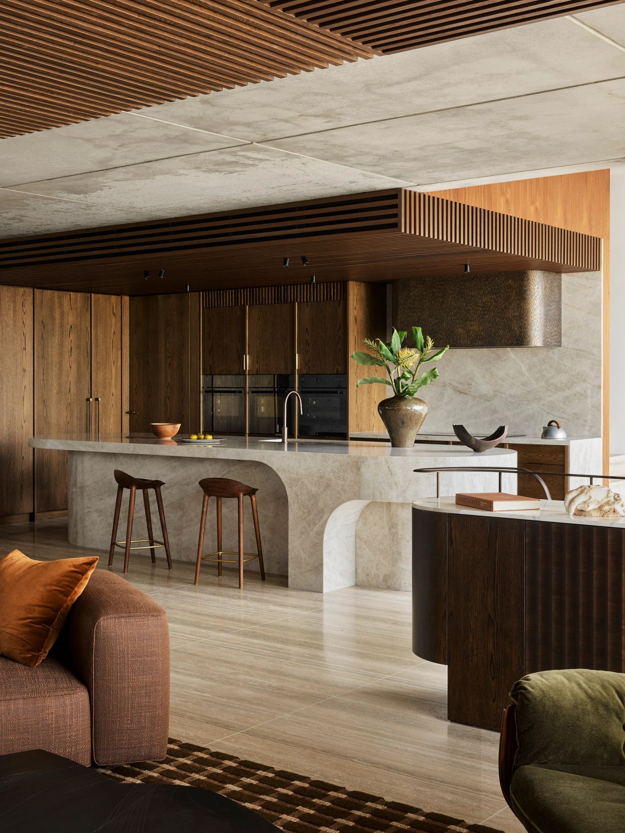Orla contemporary kitchen blending Brazilian influences with a warm brown, black, and camel palette. Features a white and beige quartzite island with timber stools, a bronze rangehood, warm timber joinery, a large ceramic vase with palms, and timber batten detailing on the concrete ceiling.