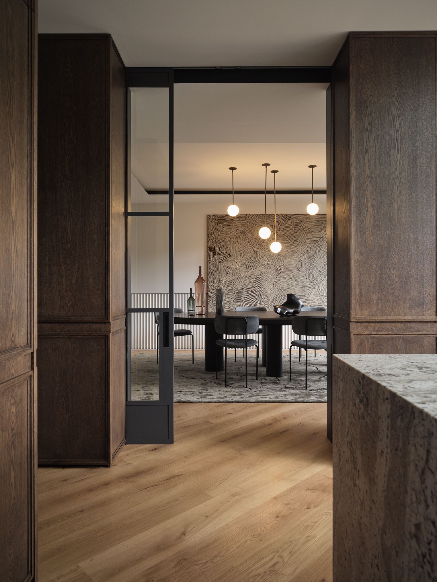 Monsieur G A view through a black steel-framed glass doorway into a dining room, framed by dark timber-panelled walls and a marble countertop edge in the foreground. Five round pendant lights hang at staggered heights above a dark round dining table with black metal-framed chairs, set on a patterned grey rug over herringbone flooring. A large abstract textured wall panel with rippling linear patterns serves as a backdrop, with decorative bottles and a sculptural object on a sideboard nearby.