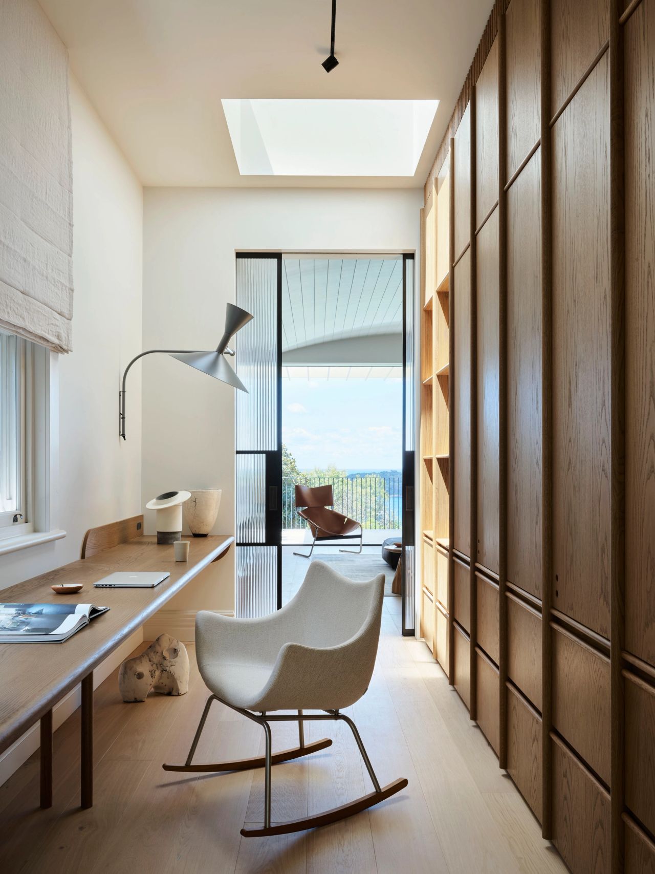 Ultramarine Narrow home office with a built-in wooden desk, cream rocking chair, sculptural arm-shaped wall lamp, and floor-to-ceiling wood cabinetry beside a glass door leading to a terrace with coastal views.