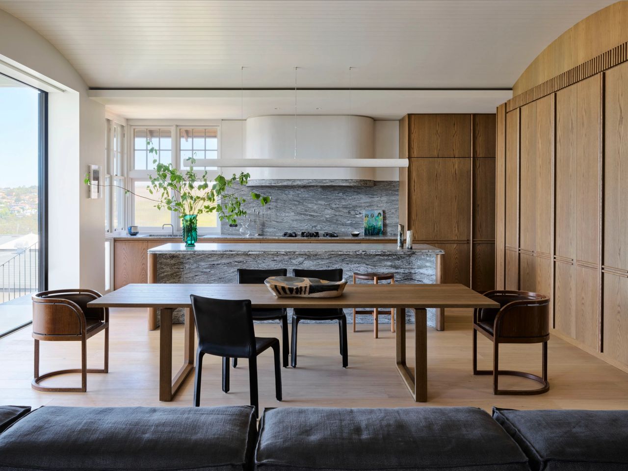 Ultramarine Open-plan dining and kitchen space with a wooden dining table, mixed black and curved wood chairs, a marble kitchen island with sculptural white range hood, and coastal views through glass doors. 