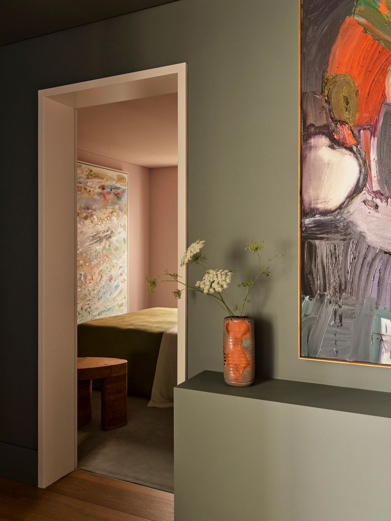 Sunset Sculptural terracotta vase with white wildflowers on a green plinth beside a bold abstract painting, with a doorway revealing a pink-hued bedroom with an olive bedspread beyond.