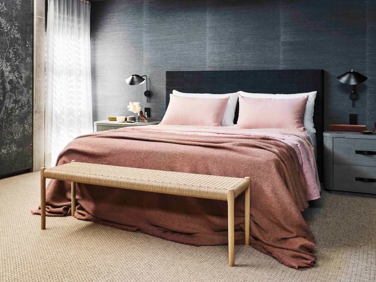 Killcare Main Bedroom with a charcoal upholstered bedhead against textured grey wallpaper, dressed with blush pink linen and a woven rush bench at the foot of the bed, flanked by black wall-mounted reading lamps