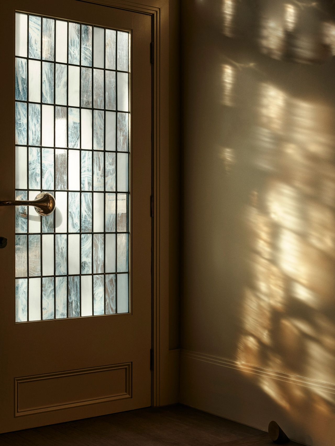 Queenie Front door with grid-patterned textured glass panels and a brass lever handle, casting dappled light and shadow patterns across the adjacent wall.