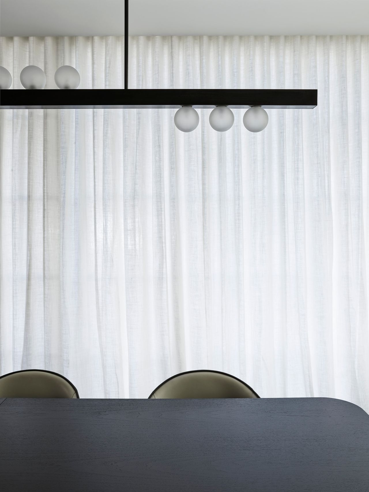 A sculptural black pendant with frosted glass globes floats above a dark dining table, sharply silhouetted against softly filtered sheer curtains.