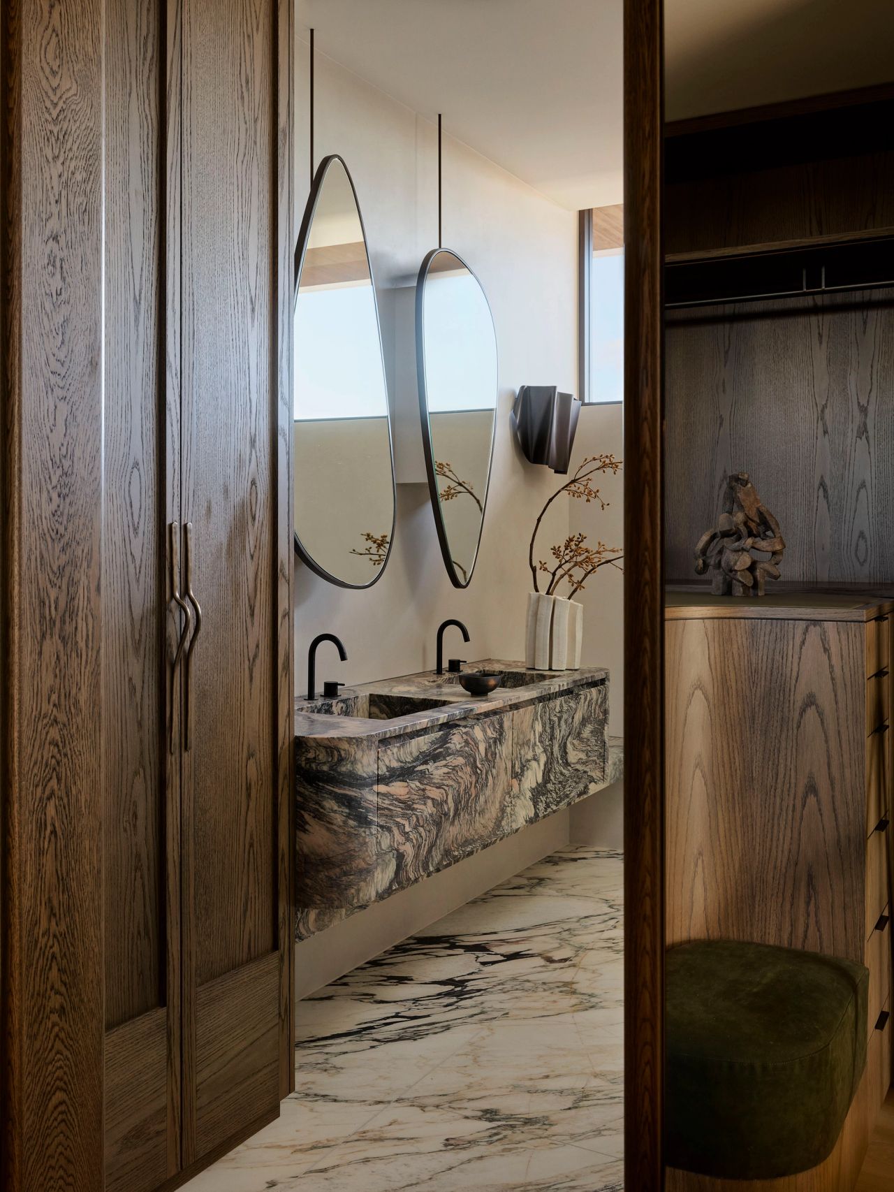 Patchwork Bathroom vanity with a dramatic veined stone floating basin, two suspended round mirrors, and black tapware, framed by dark wood cabinetry doors and dressing room shelving with a green ottoman beyond.