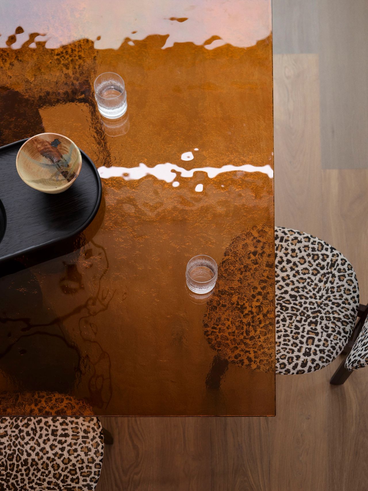 Dancing on the Ceiling Overhead view of a rust-toned glass table with two water glasses and a small ceramic bowl, surrounded by leopard-print upholstered chairs on a wooden floor.