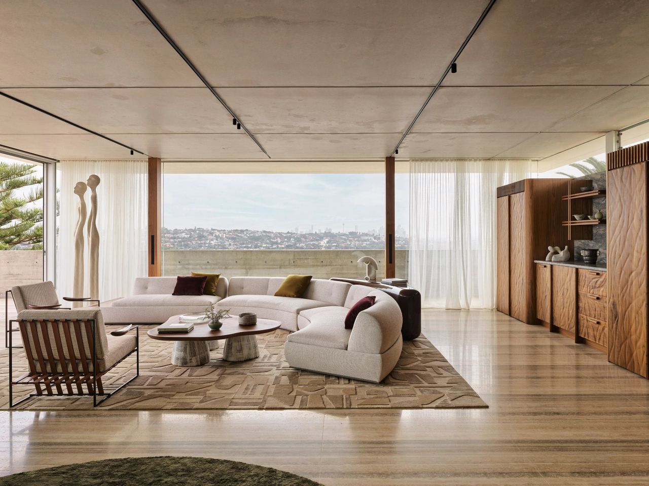 Orla contemporary open-plan living room blending Brazilian influences. A walnut coffee table with cream-glazed ceramic legs sits beside a Brazilian-style occasional chair with beige upholstery and timber arm detailing. A cream curved modular lounge sits on a geometric-patterned rug, with expansive views over Sydney's eastern suburbs and CBD, and walnut bar joinery featuring custom curved texture detailing.