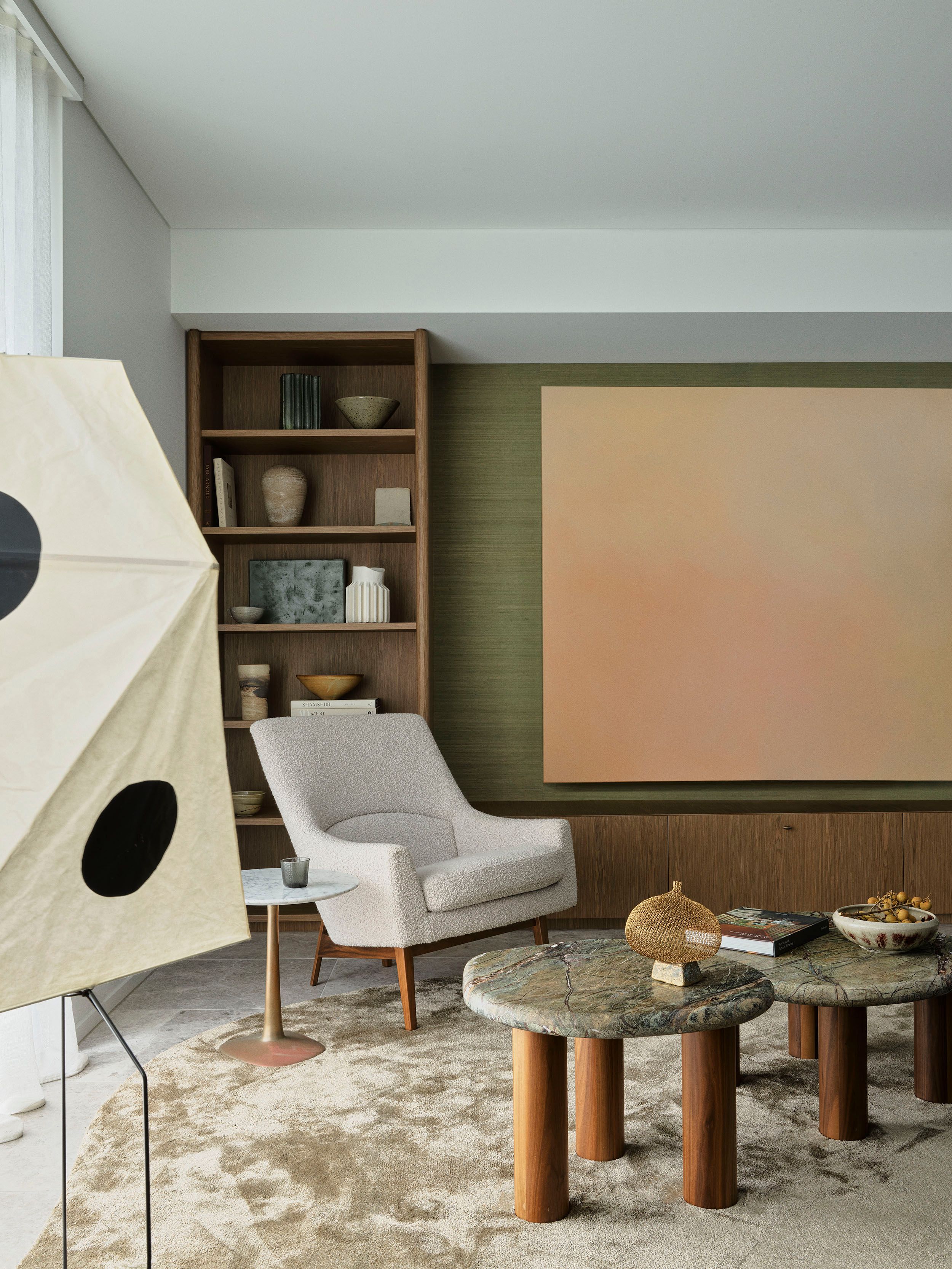Sculptural stone tables, warm timber joinery and a textured boucle armchair sit against olive walls and large-scale artwork.