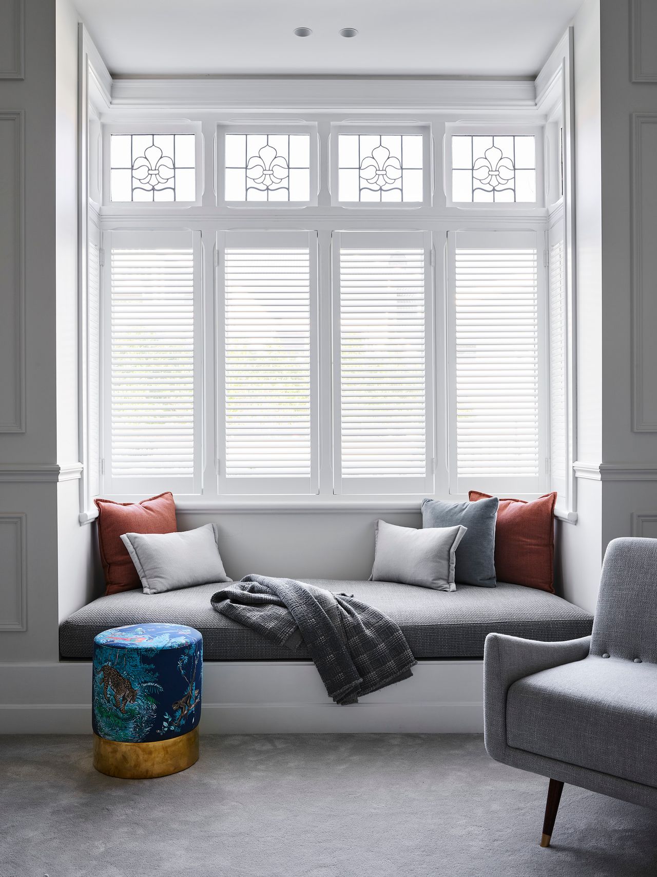 Elegant window seat with grey upholstery and rust cushions beneath decorative leadlight windows and white plantation shutters.