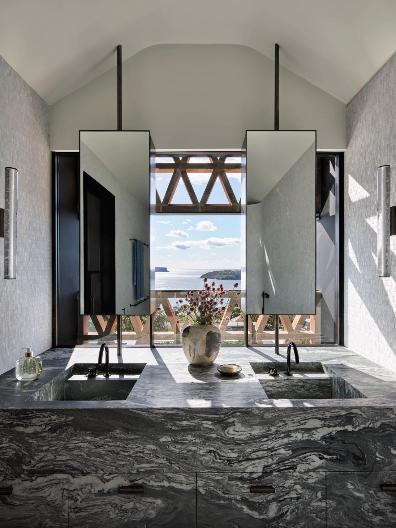 Ultramarine Double vanity in dark veined marble with two suspended angled mirrors, integrated basins, and black tapware, framed by a large window with a decorative timber truss and coastal ocean view.
