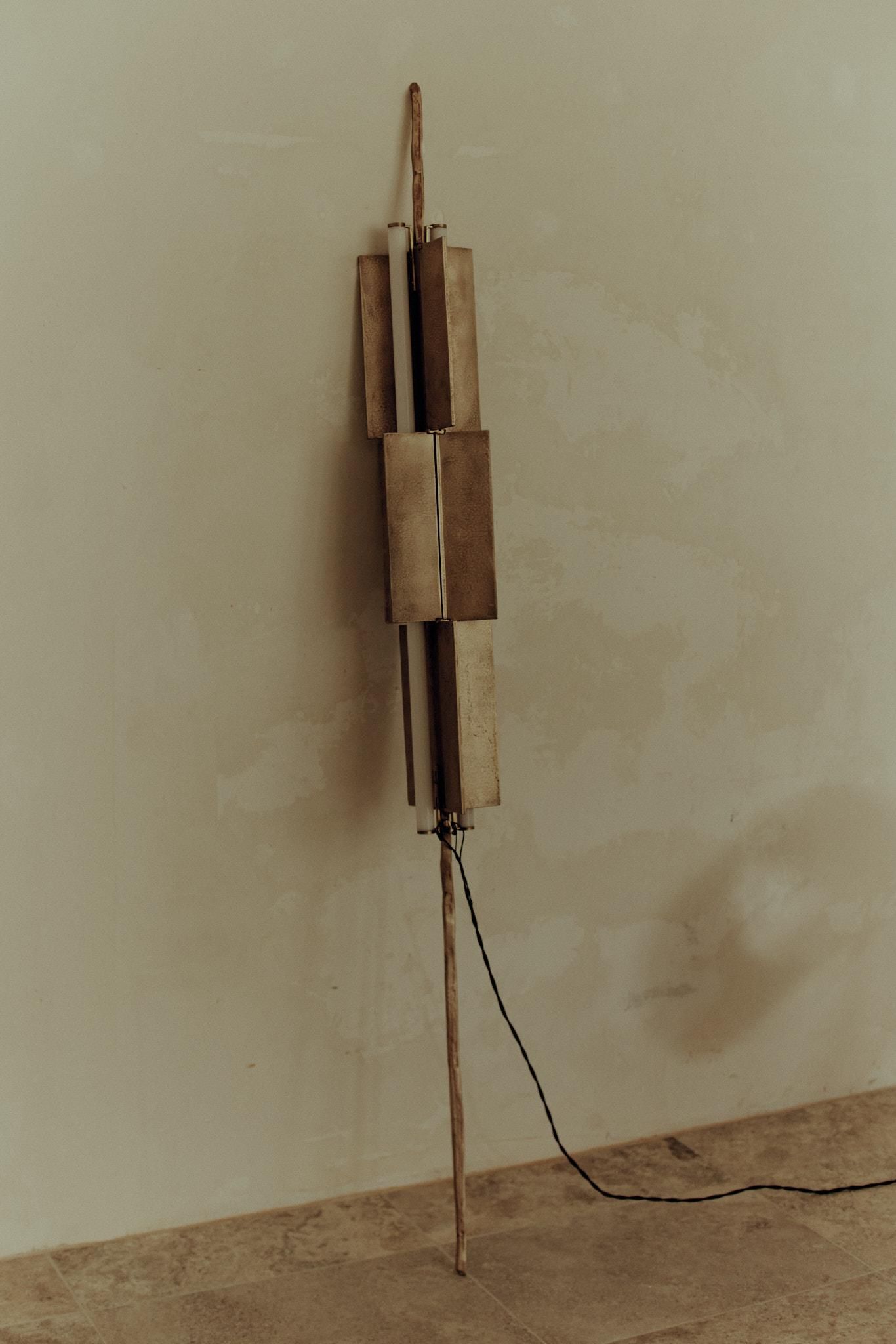 MTT-1 floor lamp by Linda Valentic, a sculptural composition of layered metal forms and a branch-like stem against a softly textured plaster wall.