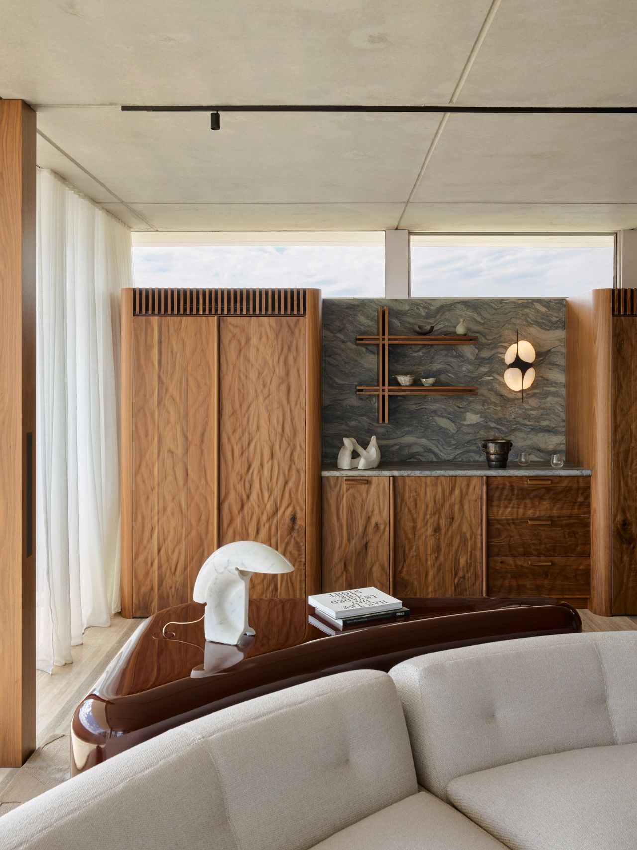  Orla contemporary living room blending Brazilian influences. Detail of a cream curved modular lounge sits on a geometric-patterned rug, with a wine coloured high-gloss table. behind is a wall-mounted walnut timber shelf with a crossed intersection detail, set against a blue-and-beige stone wall and styled with a ceramic bowl