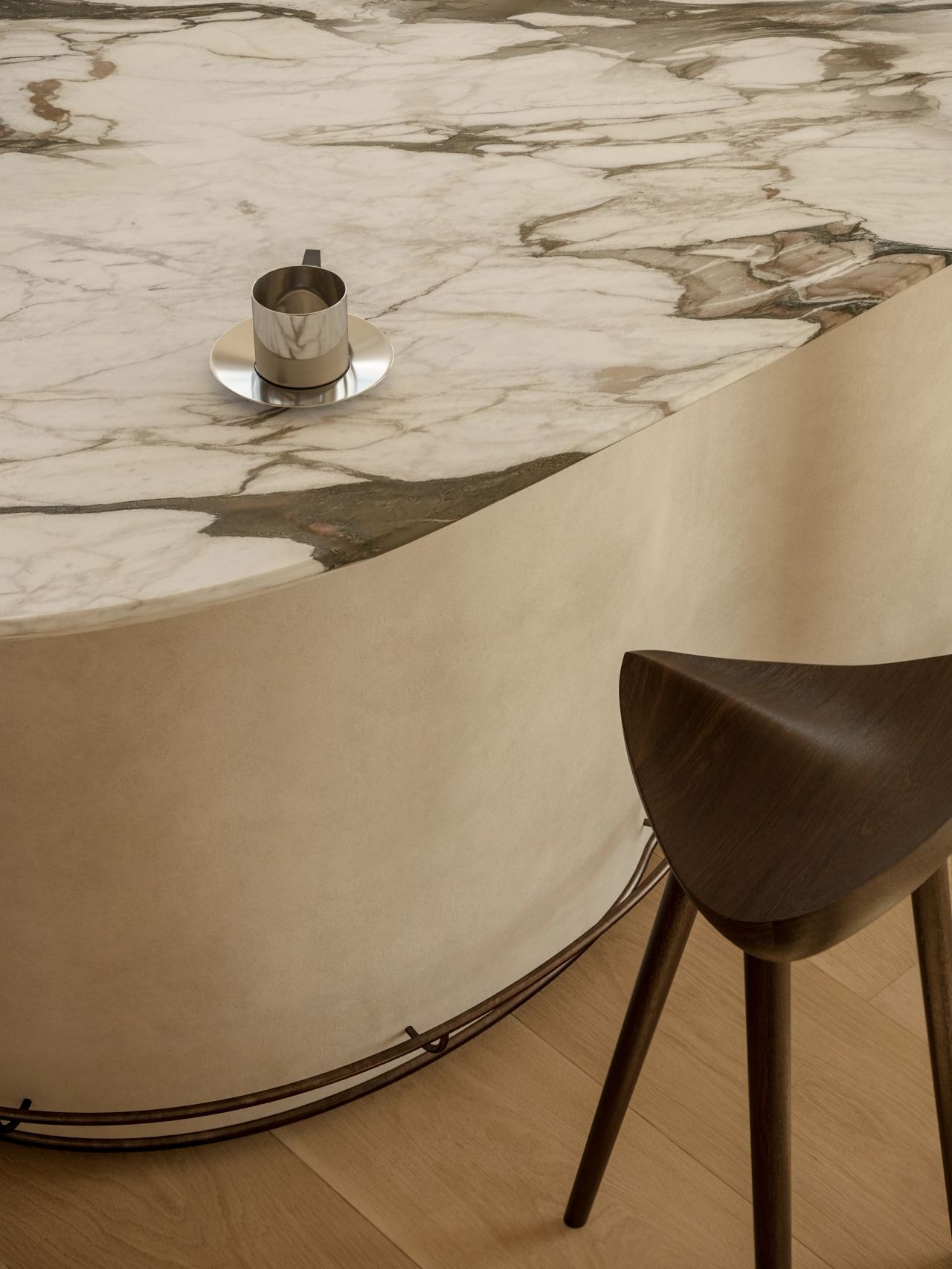 Lady M Close-up detail of a curved kitchen island with dramatic gold-and-white veined marble top and plaster-finish base, styled with a polished metal cup and saucer. A sculptural dark timber bar stool with a brass footrail sits alongside