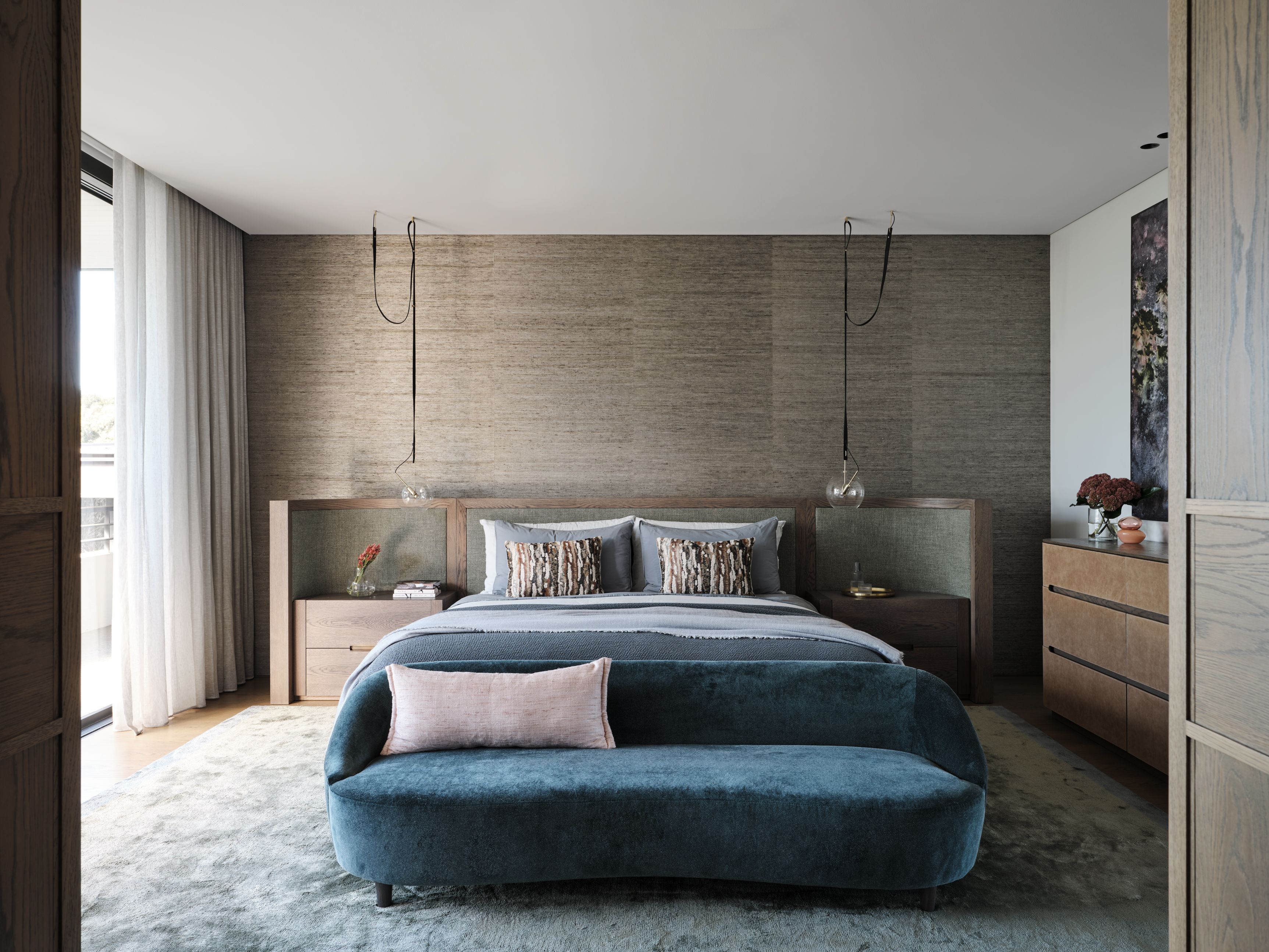 Sophisticated contemporary bedroom layering warm timber, textured wall covering and soft blue-grey textiles, anchoring by a sculptural velvet bench.