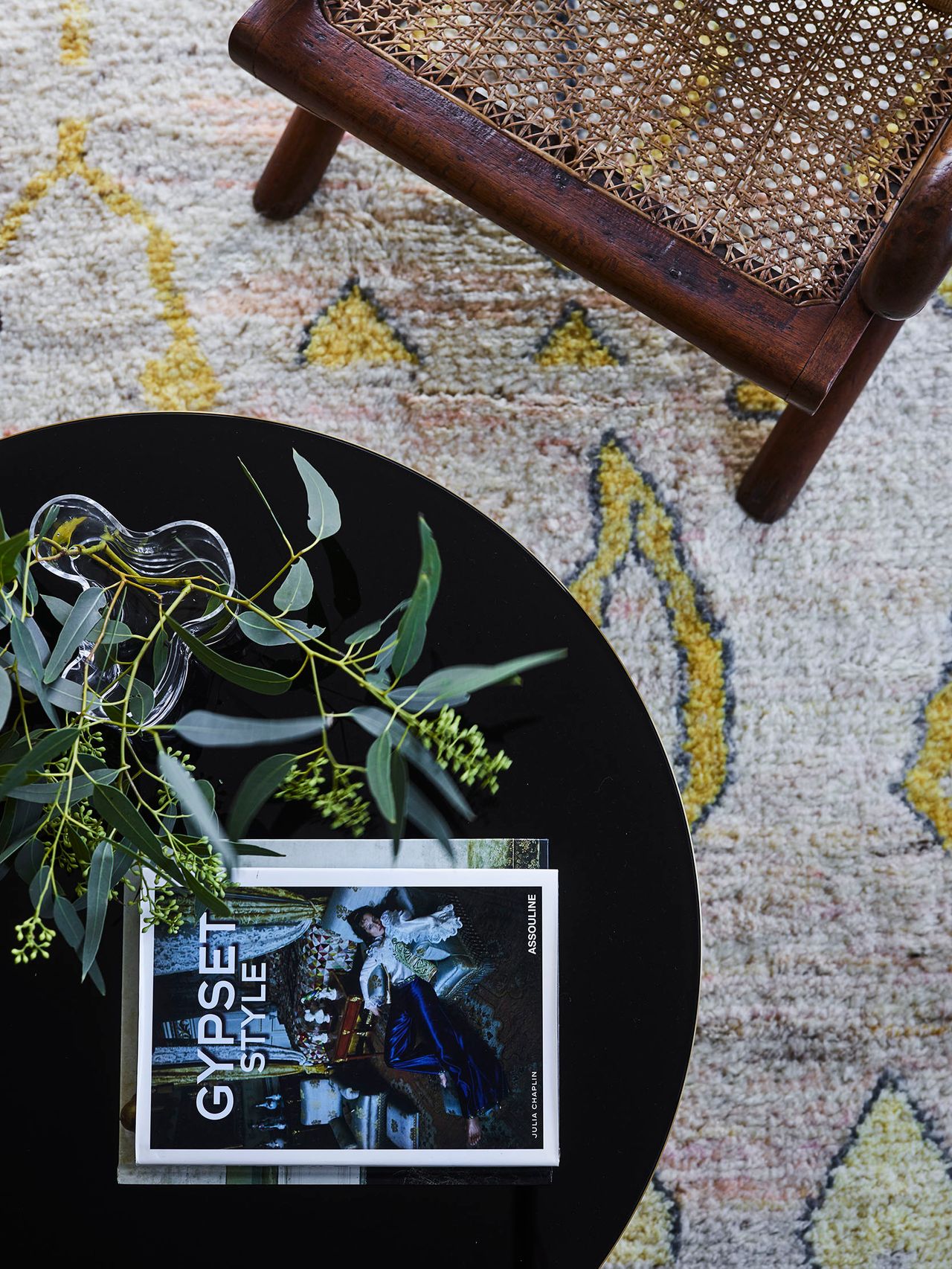 Killcare Overhead view of a round black side table with a glass bud vase of eucalyptus and a coffee table book, beside a cane-seated timber stool on a patterned cream and yellow rug