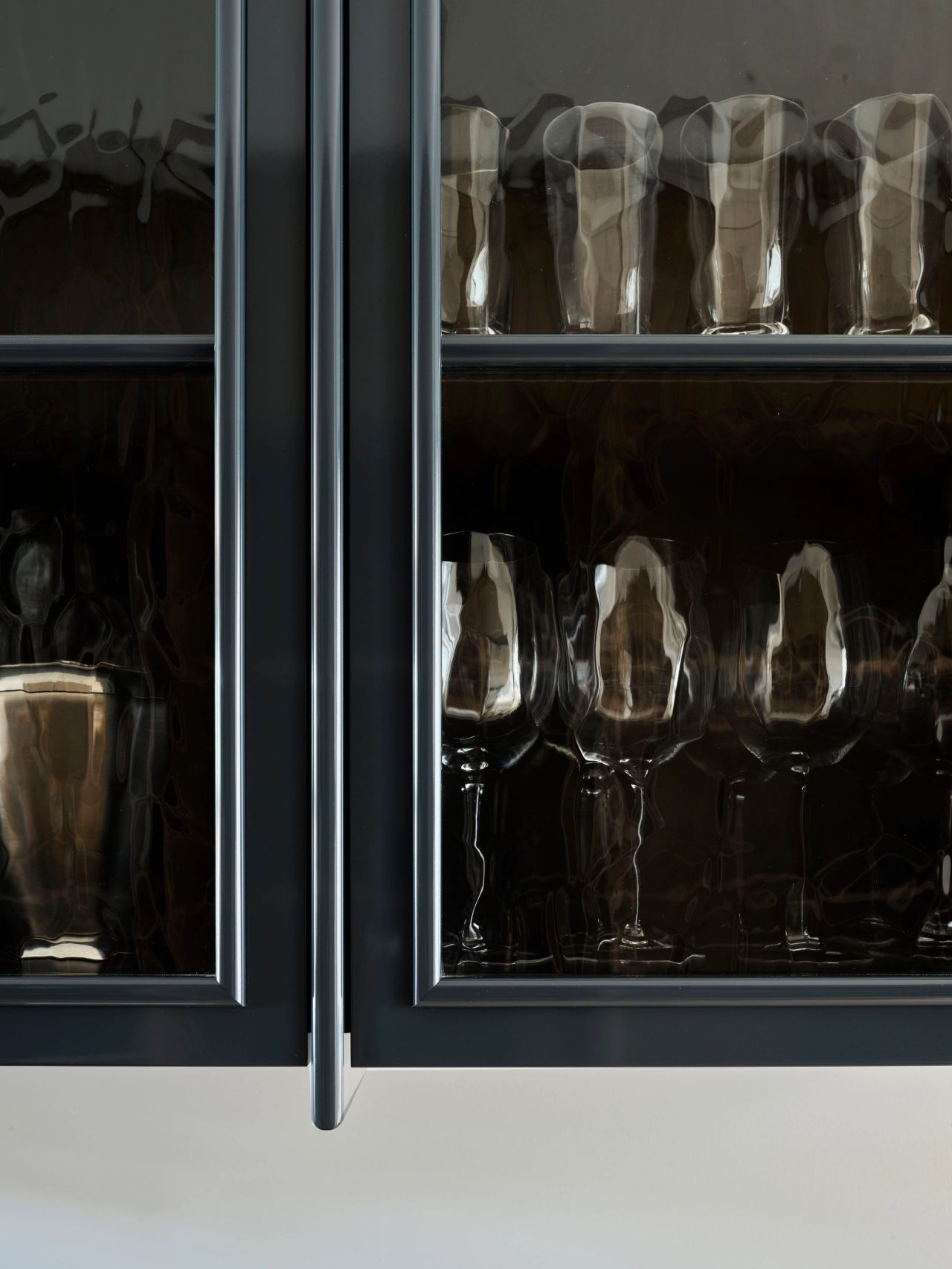 Ultramarine Close-up of dark navy glass-front cabinets with slim brass handles, displaying rows of textured glassware including tumblers and stemmed wine glasses