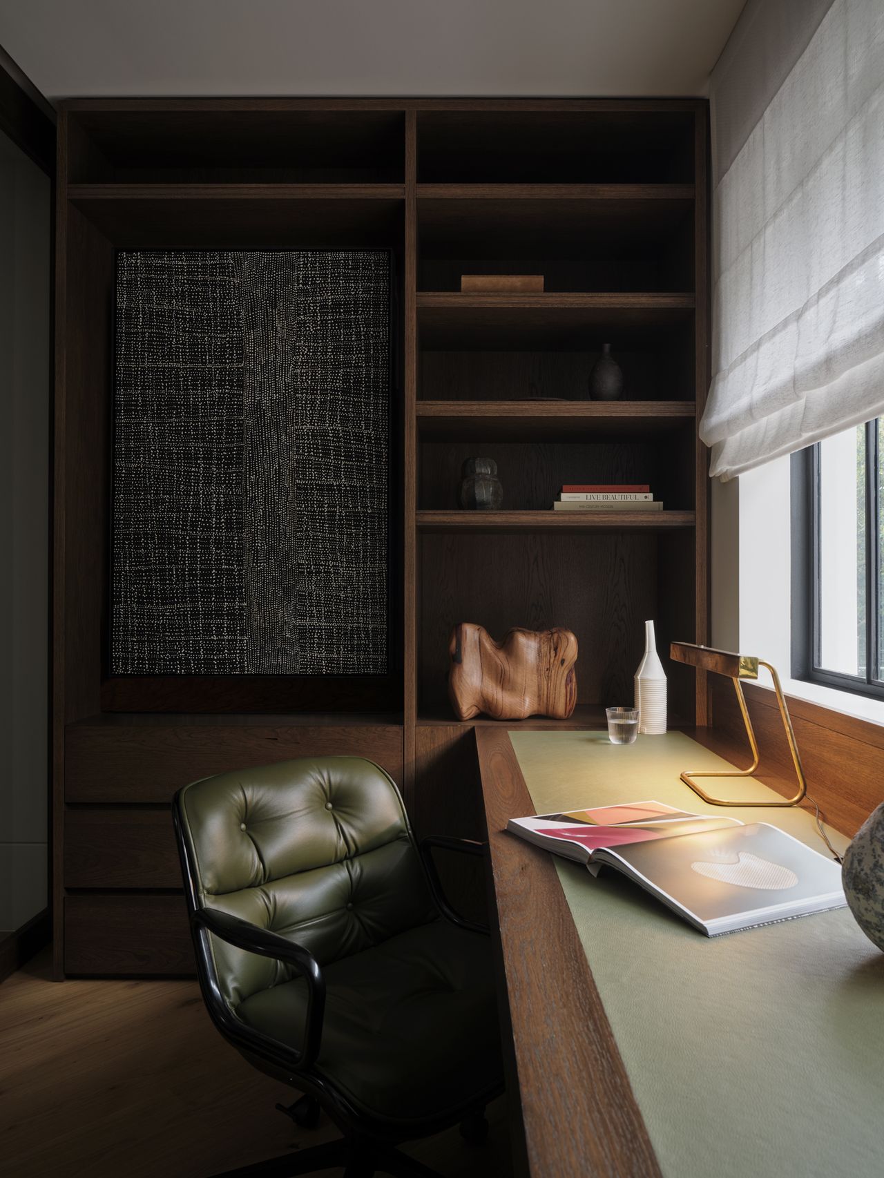 Monsieur G Study with built-in dark timber joinery and open shelving displaying books and a carved wood sculpture, an olive leather tufted desk chair, a green leather-topped desk with brass lamp, and a sheer roman blind at the window