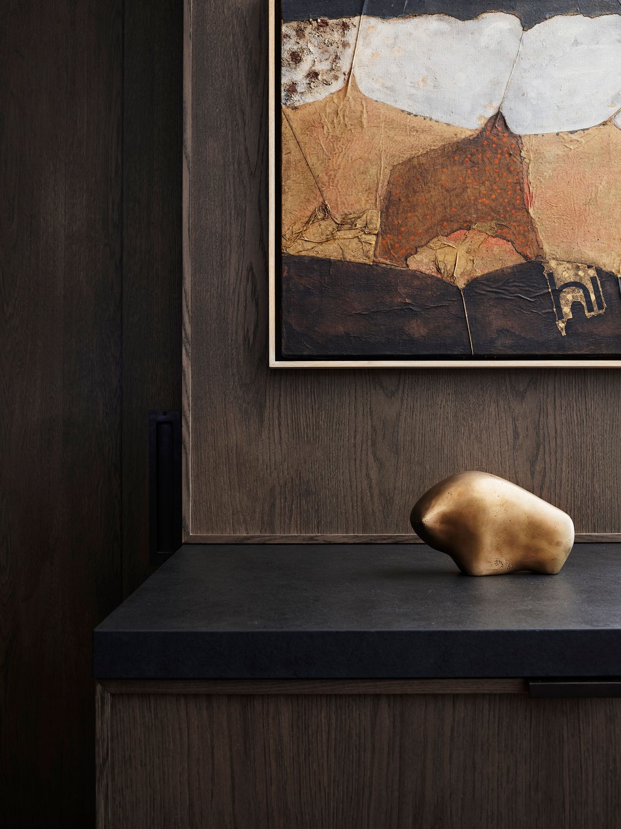 A bronze, sculptural object sits beneath a richly layered textured abstract artwork, set against dark timber joinery in this moody coastal interior by Decus.
