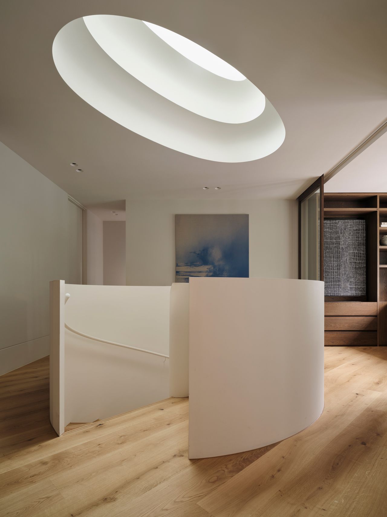 Monsieur G A minimalist interior landing featuring a large oval recessed skylight with concentric stepped rings set into the ceiling, echoing the sweeping curved white balustrade of a spiral staircase below. A framed abstract blue and white artwork hangs on the wall behind, with light oak flooring and a glimpse of a wood-panelled hallway to the right