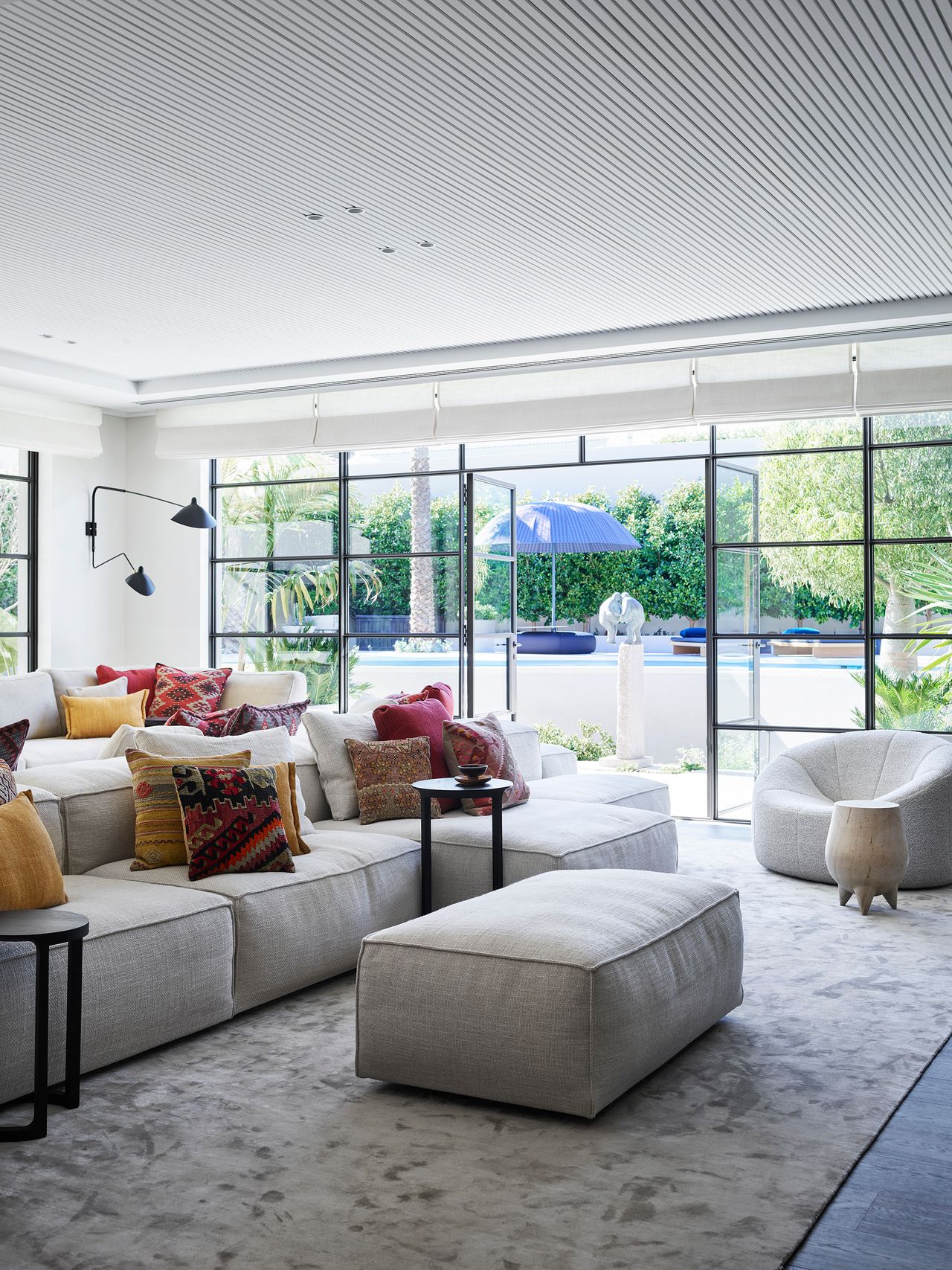 Generous neutral seating layered with richly patterned cushions creates a relaxed, colourful living space, opening through steel-framed doors to the pool and lush garden beyond.
