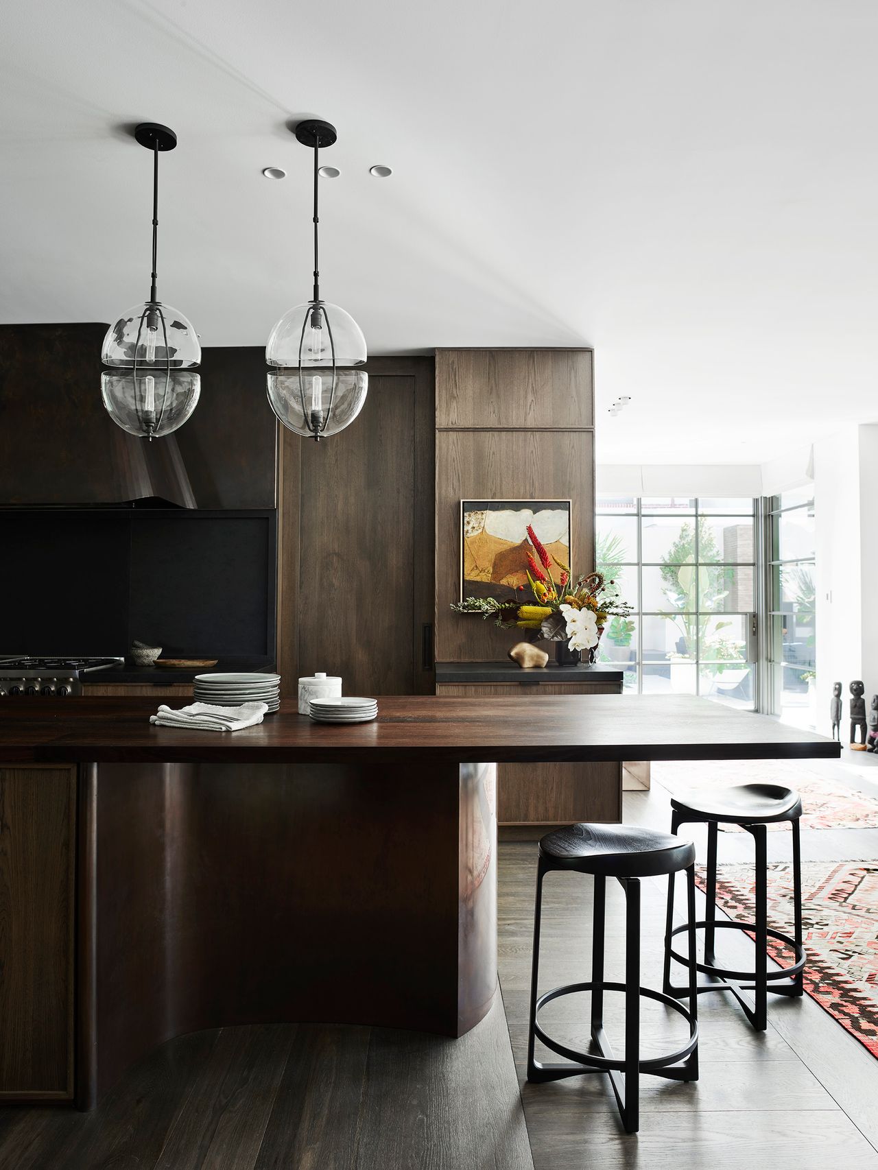 Contemporary kitchen with dark timber cabinetry, a sculptural bronze island, black bar stools and glass pendant lights, opening to a bright garden terrace.