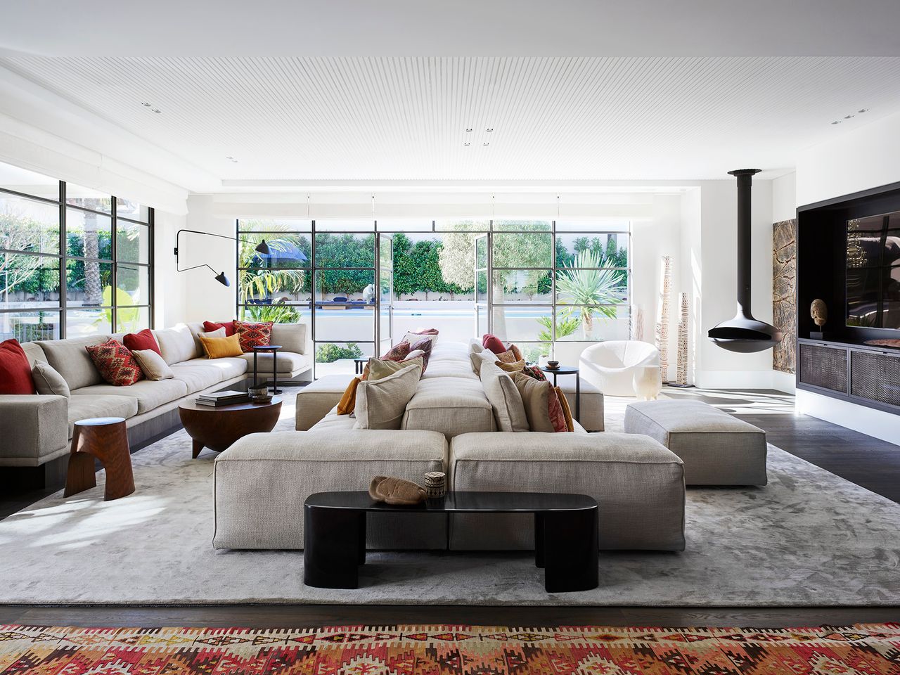 Light-filled contemporary living room with large neutral sofas, colourful cushions, a suspended black fireplace and steel-framed doors opening to a lush garden and pool.