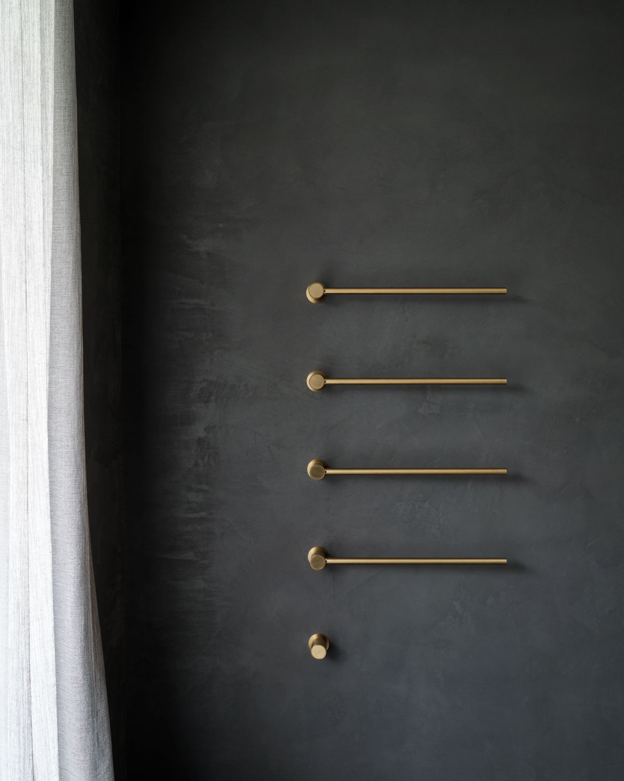 Minimalist bathroom detail with brushed brass towel rails mounted on a dark charcoal textured wall beside sheer curtains.