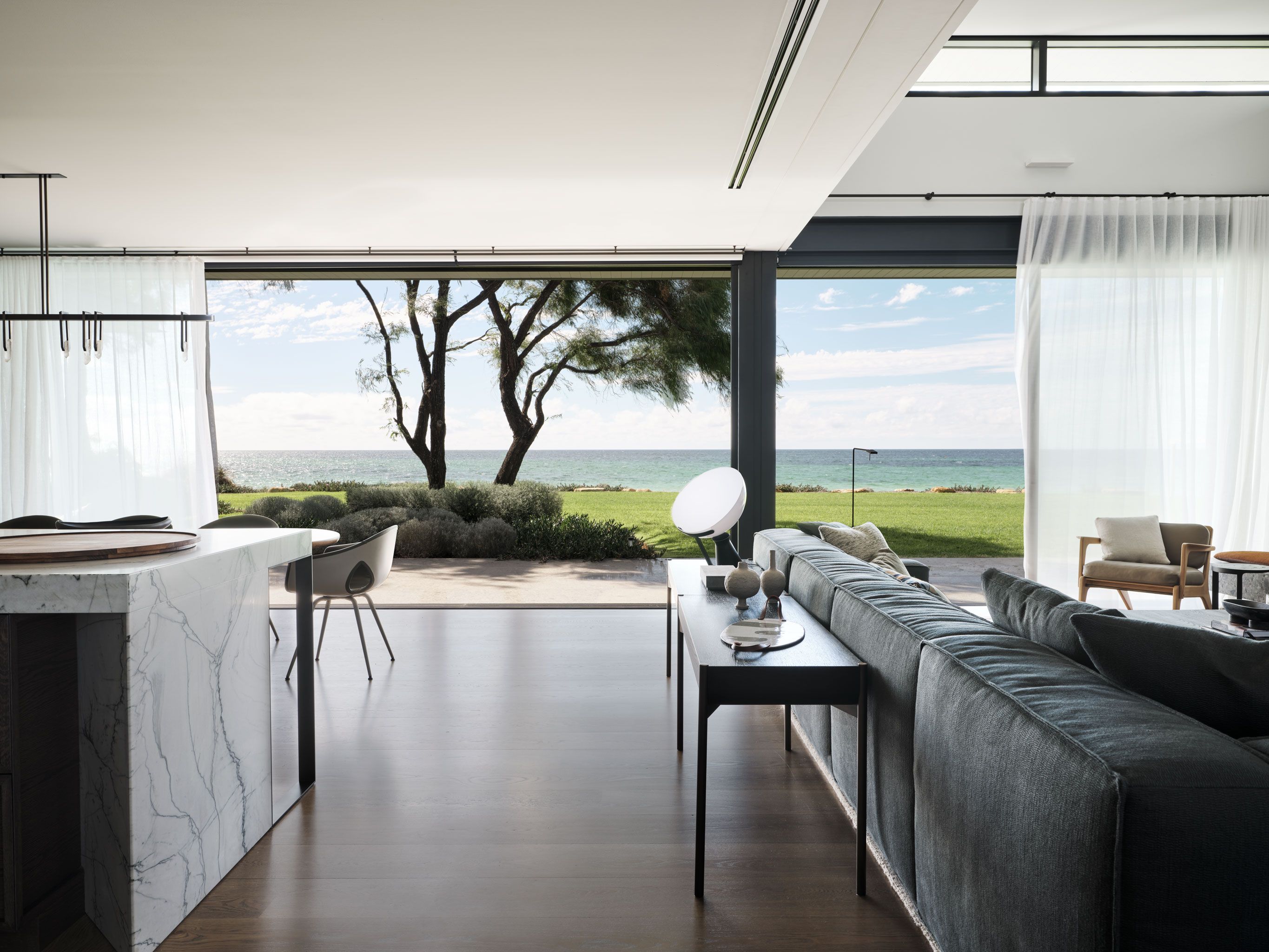 Contemporary open-plan coastal living room and kitchen with marble island, charcoal sofa and expansive ocean views.