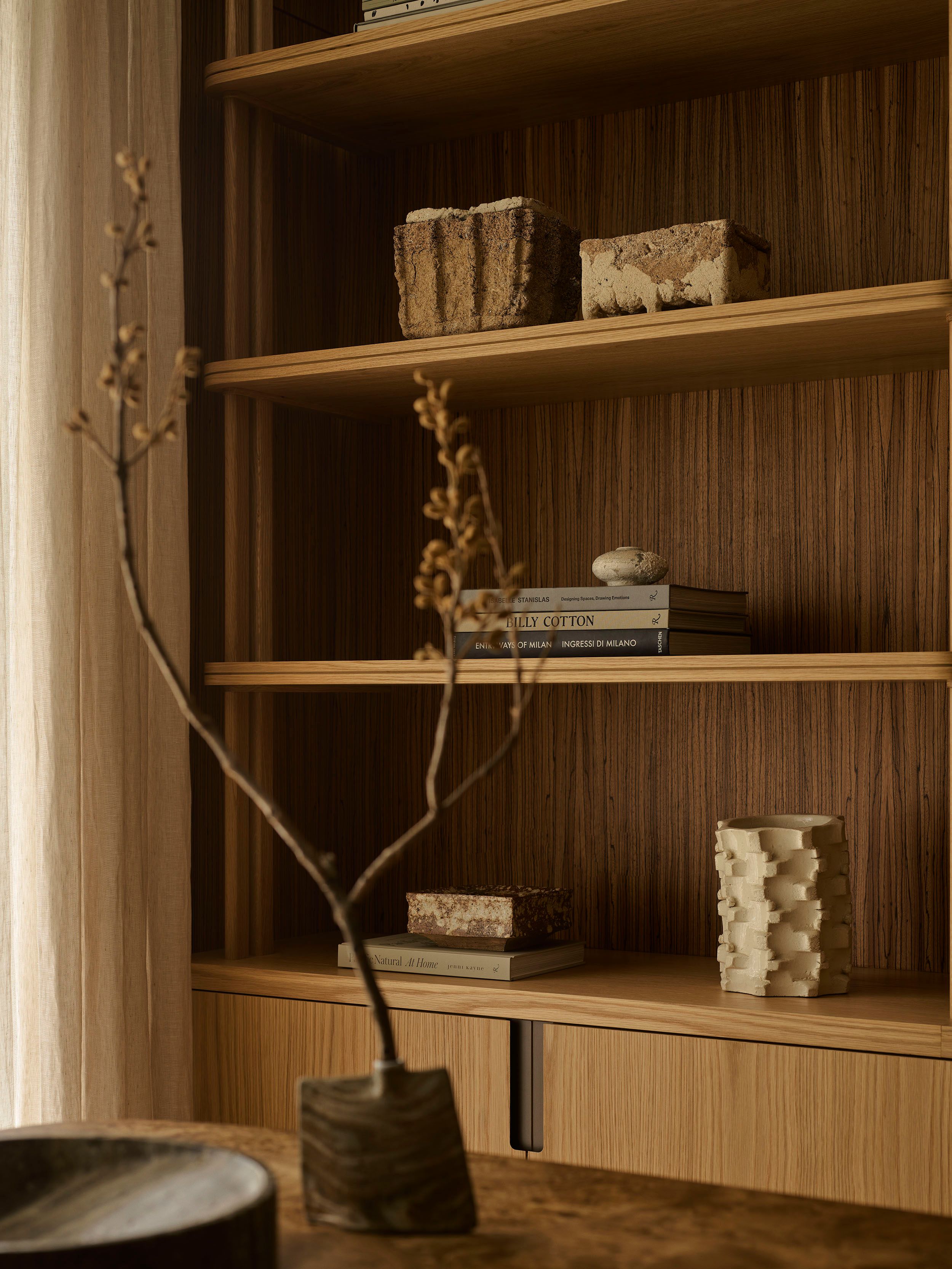 Patchwork Styled wooden bookshelf with textured stone objects, stacked coffee-table books, and a sculptural ceramic vase, softly lit with a sheer curtain and dried branch in the foreground.