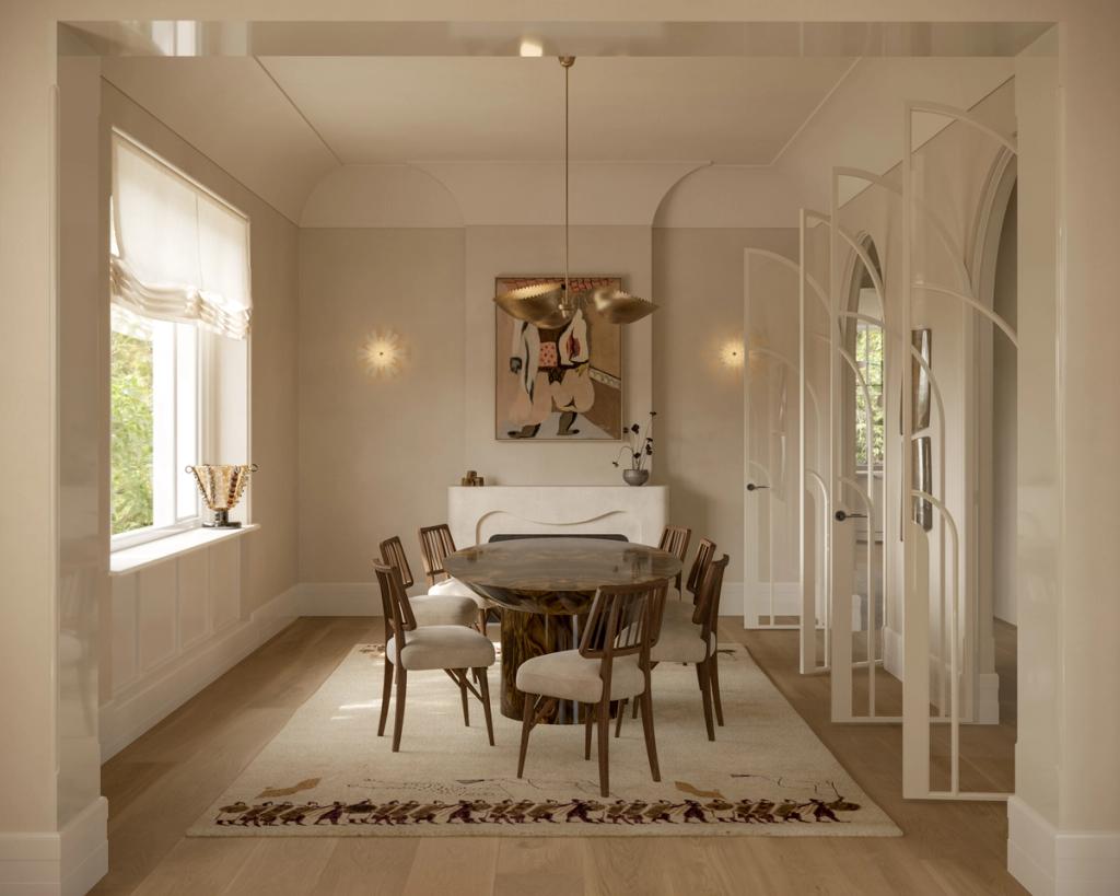 Lady M Dining room with a round burl-wood table and upholstered timber chairs on a patterned rug, a figurative artwork above a plaster mantel, starburst wall sconces, arched panelled doors, and Roman blinds at the window