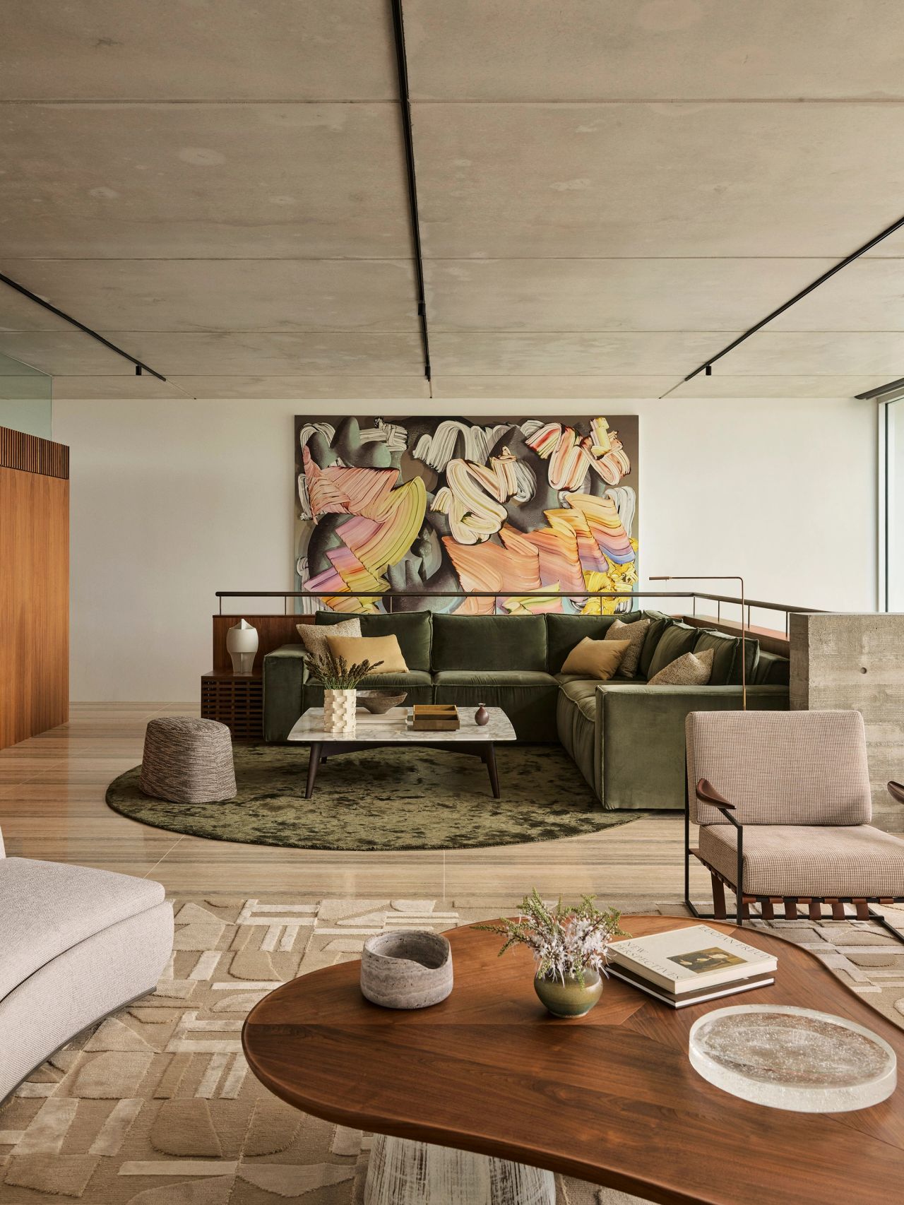Orla contemporary open-plan living room blending Brazilian influences, with a forest-green sofa on a forest-green silk rug and a large multicoloured artwork above the stairwell behind. In the foreground, a coffee table with a walnut timber top and cream-glazed ceramic legs sits beside a Brazilian-style occasional chair with beige upholstery and timber arm detailing.