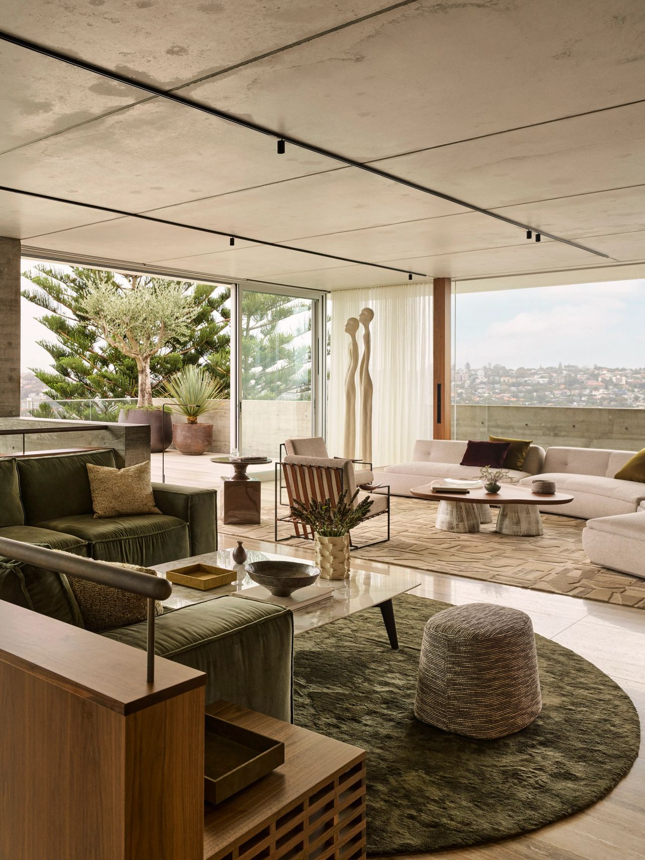 Orla contemporary open-plan living room blending Brazilian influences, with a forest-green sofa on a round forest-green silk rug. In the background, a coffee table with a walnut timber top and cream-glazed ceramic legs sits beside a Brazilian-style occasional chair with beige upholstery and timber arm detailing.  Cream-coloured curved modular lounge. Expensive views over Sydney’s eastern suburbs. 