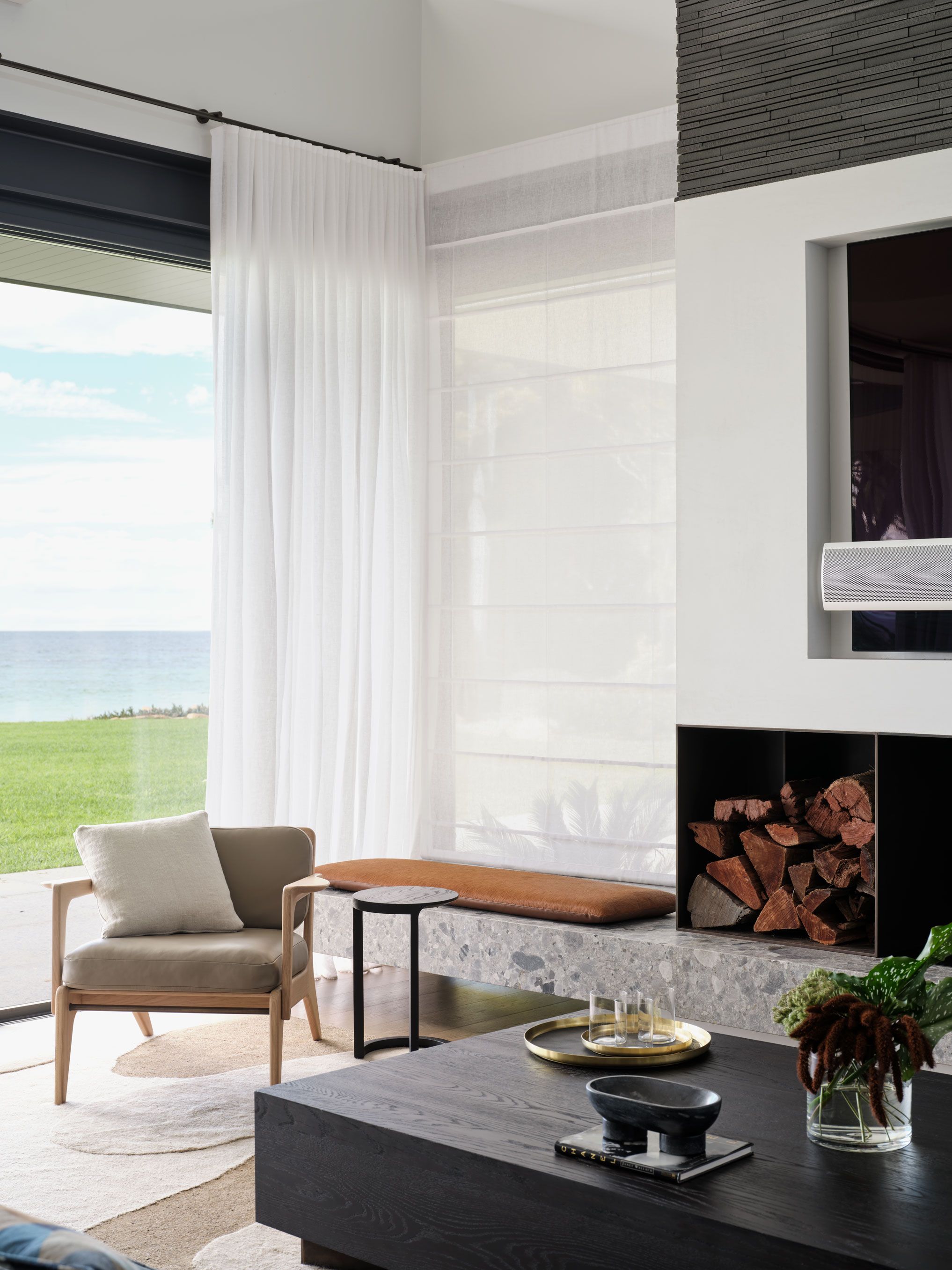 Light-filled coastal living room overlooking the ocean, featuring soft neutral furnishings, a stone hearth and warm natural timber details.