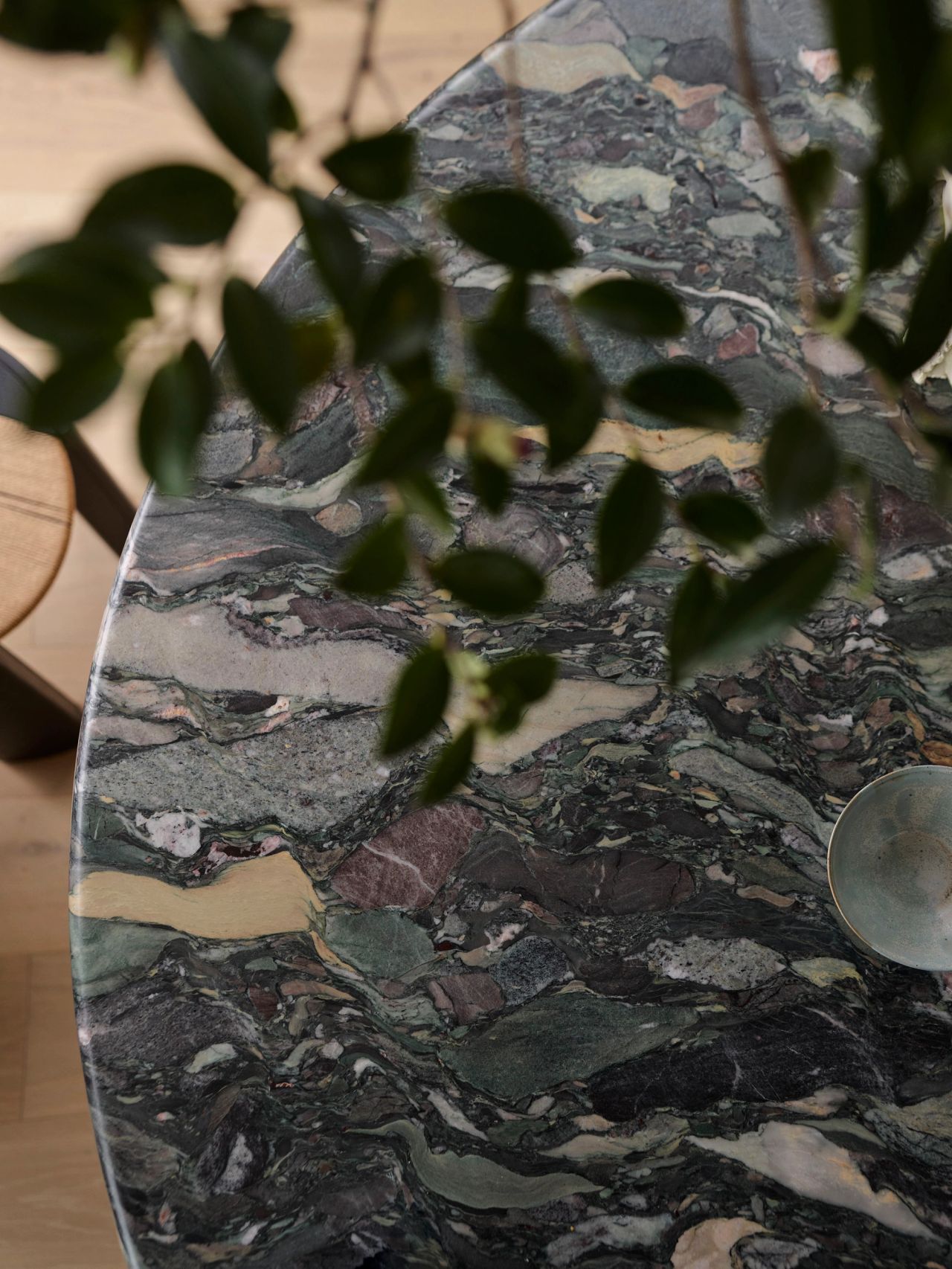 Patchwork Close-up of a round table with dramatic green, grey, and gold breccia marble top, partially obscured by out-of-focus dark green leaves in the foreground, with a small brass dish visible at the edge.