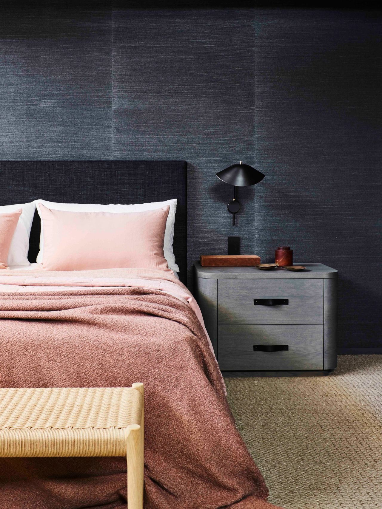 Killcare Bedroom detail with a curved grey timber bedside table and black wall lamp against textured charcoal wallpaper, styled with blush pink bedding and a woven rush bench