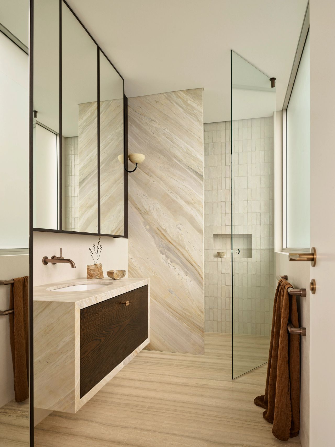 Orla ensuite with warm beige-grey stone shower walls and a matching vanity featuring a timber inlay drawer. Travertine floors, cream handmade shower wall tiles, aged bronze hardware, and a rust-coloured towel complete the space.\