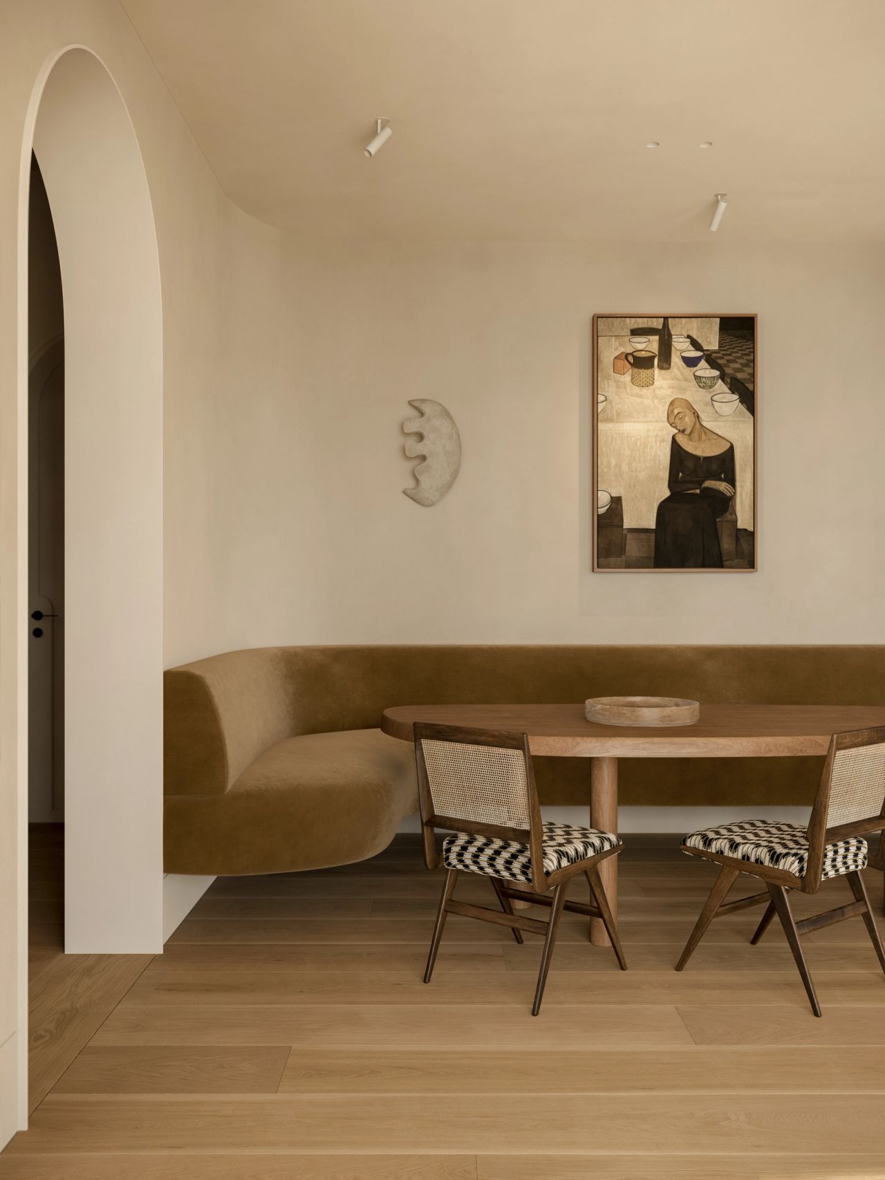 Lady M Breakfast nook with a curved olive-green velvet banquette wrapping an oval timber table, paired with cane-back chairs upholstered in black-and-white check. A figurative portrait painting and sculptural moon-shaped wall piece hang above, with an arched doorway to the left