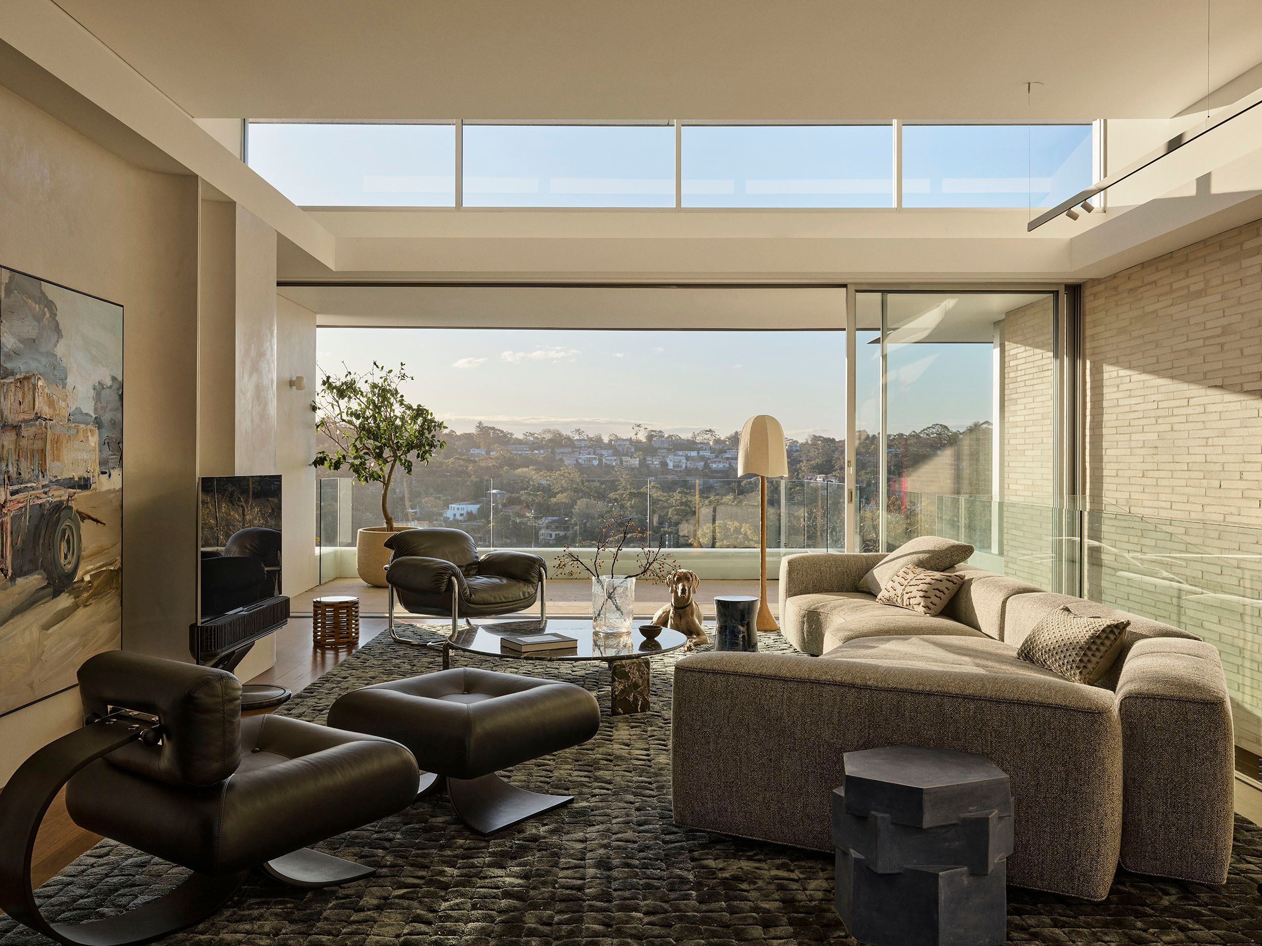 Sunset Living room with dark leather lounge chairs on a chunky woven rug, a beige sectional sofa with patterned cushions, a sculptural black side table, and glass walls opening onto sweeping treetop and hillside views.