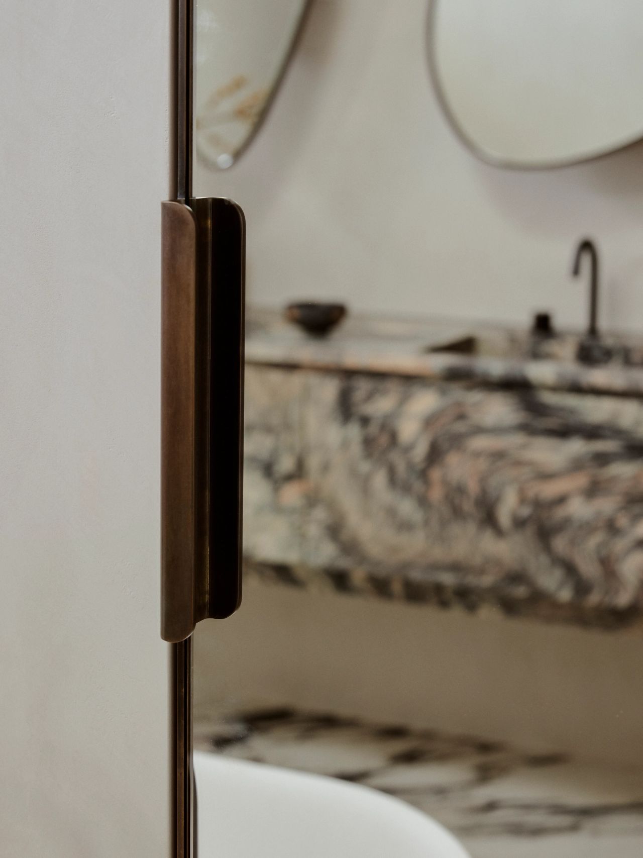 Patchwork Close-up of a bronze shower door handle in the foreground, with a blurred marble vanity, round mirror, and black tapware visible behind the glass