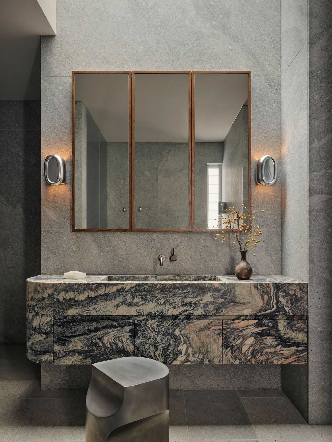 Sunset Bathroom vanity in dramatic swirling marble beneath a tri-panel wood-framed mirror, flanked by oval brushed-metal sconces, with a small vase of yellow blossoms and a sculptural stone stool in front