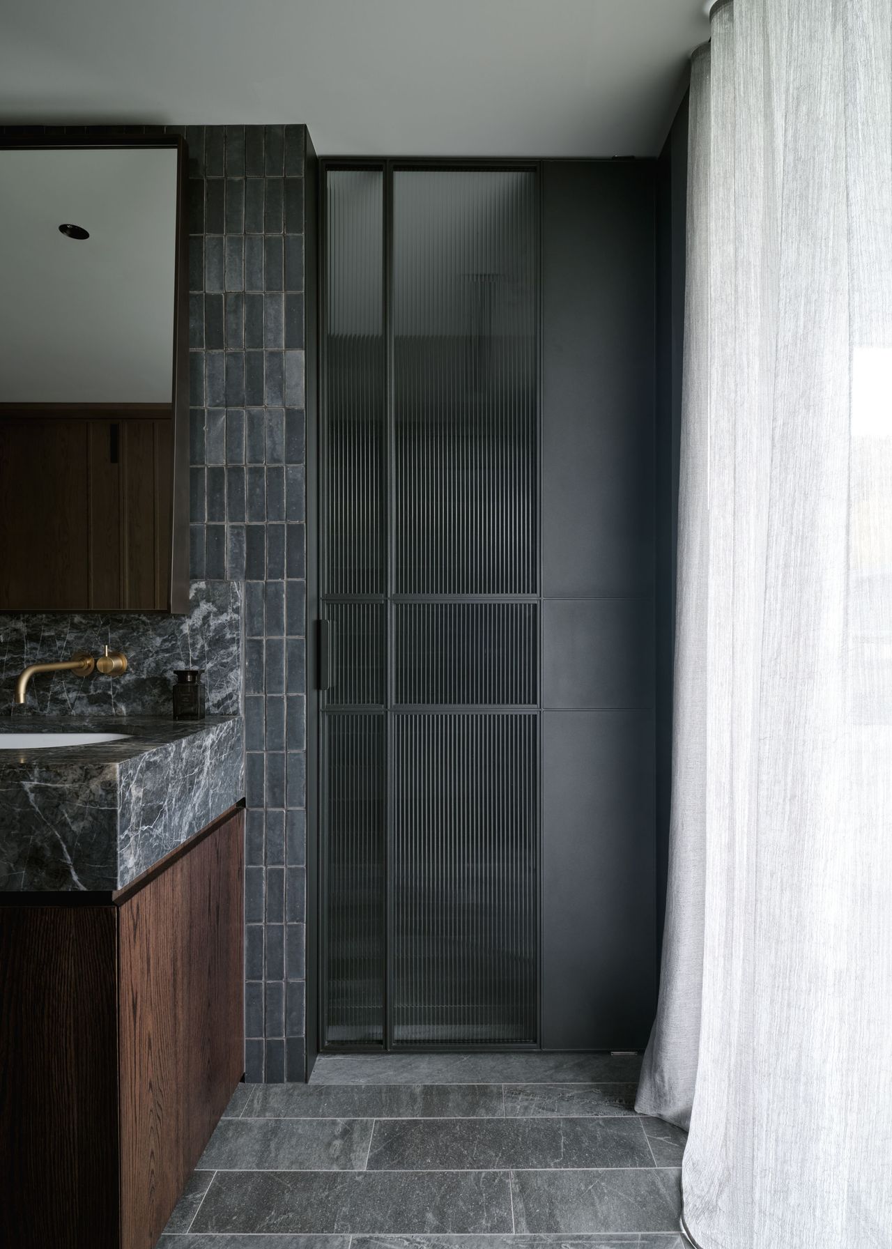 A layered bathroom composition of dark timber, veined stone and hand-glazed tiles, softened by fluted glass and floor-to-ceiling sheer drapery.