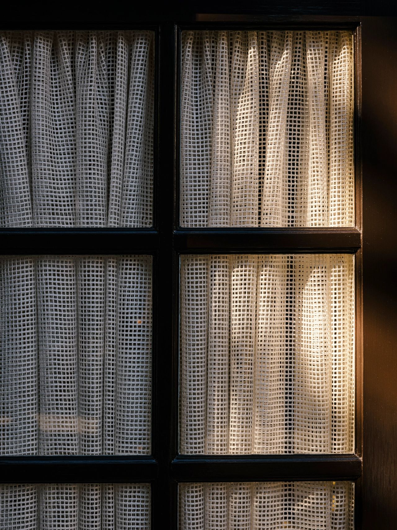 Queenie Close-up of a black-framed four-pane window with sheer, open-weave textured curtains, sunlight streaming through one panel