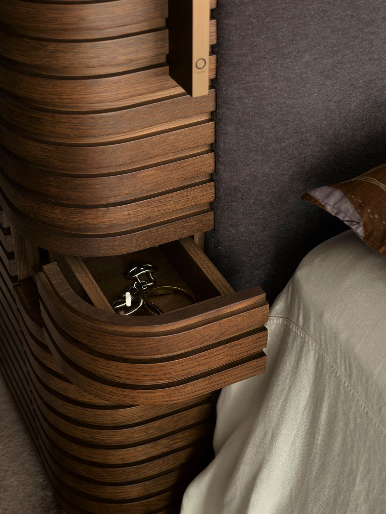 Sunset Close-up of a curved, ribbed wooden bedside unit with an open drawer revealing bracelets, beside a purple upholstered headboard and neutral bedding