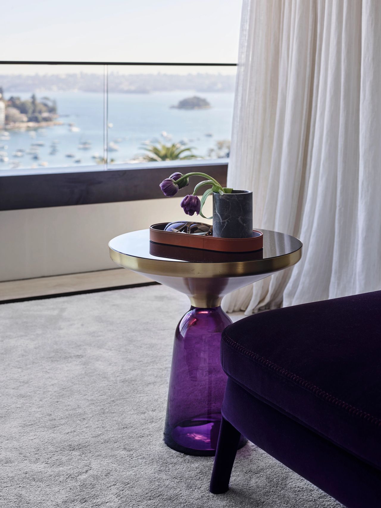 Sculptural purple glass side table beside a velvet chaise, styled with purple flowers and overlooking a sunlit harbour.
